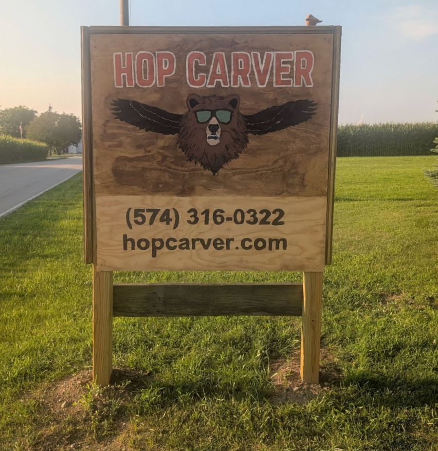 Outdoor wooden sign for Hop Carver with a bear wearing sunglasses and wings, phone number, and website.