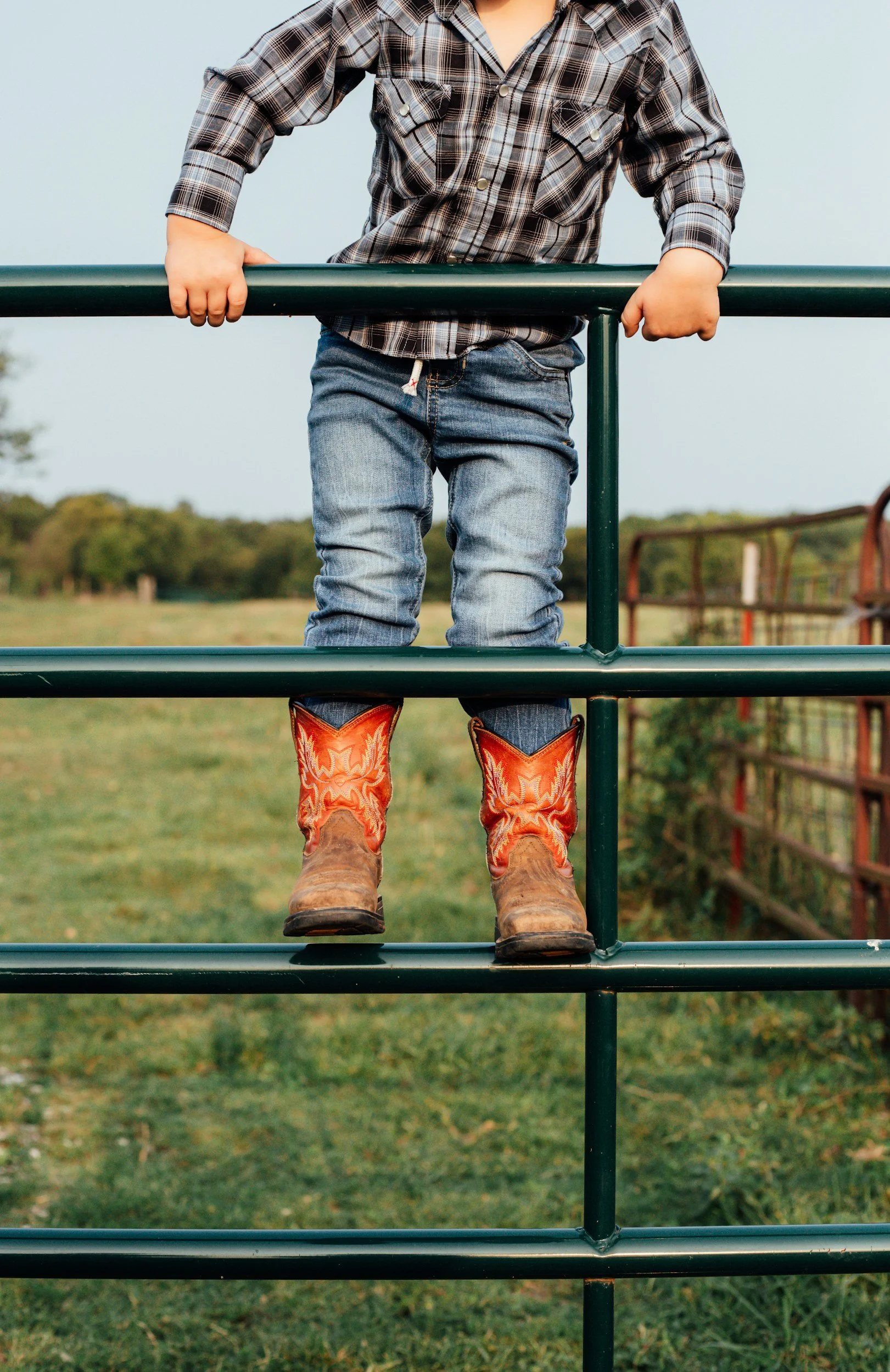 A child standing on a green metal fence, wearing a plaid shirt, jeans, and cowboy boots, outdoors in a rural setting.