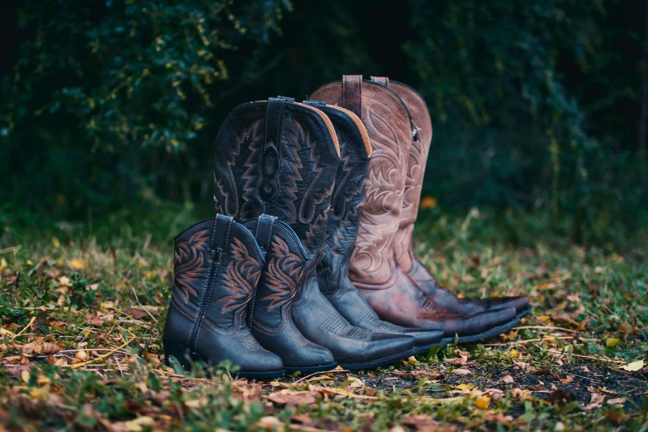 Three pairs of cowboy boots, patterned with embroidery, lined up on a grassy ground amid fallen leaves with greenery in the background.