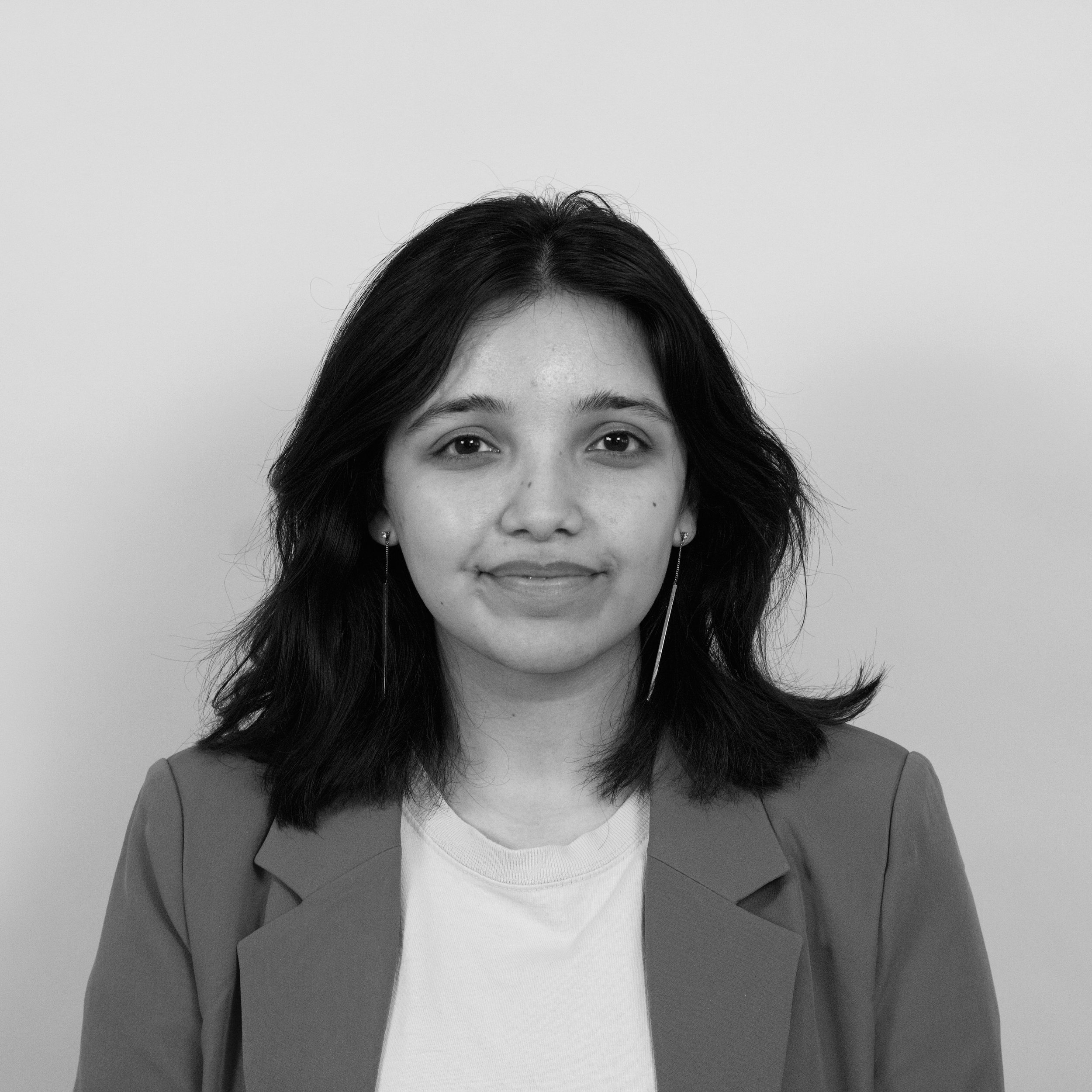 A black-and-white headshot of Khadija Alam smiling at the camera. She has shoulder-length black hair and is wearing a blazer, a t-shirt, and long earrings.