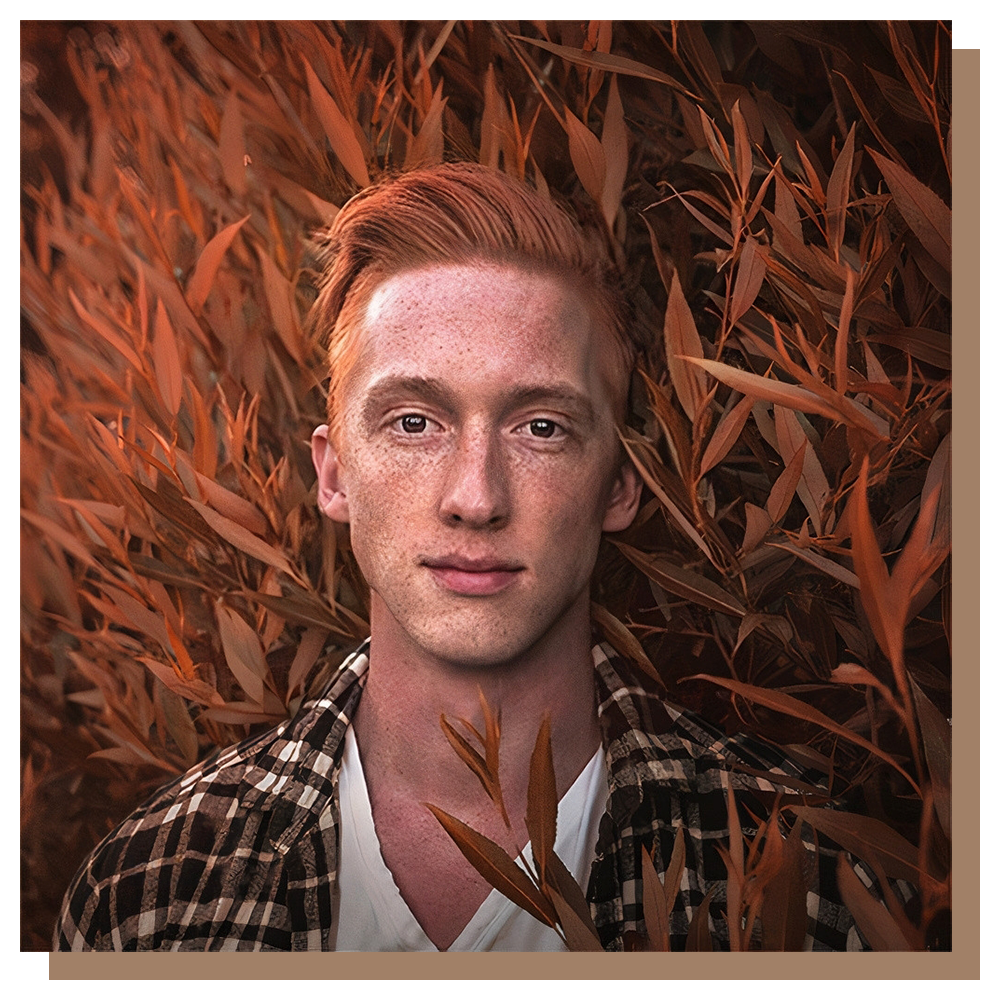 Young man with light skin and red hair, wearing a plaid shirt, standing among tall, orange-colored plants or tall grass.