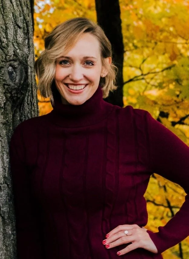 A woman with blonde hair and a cheerful smile, wearing a maroon turtleneck sweater, standing outdoors in front of a tree with autumn yellow leaves.