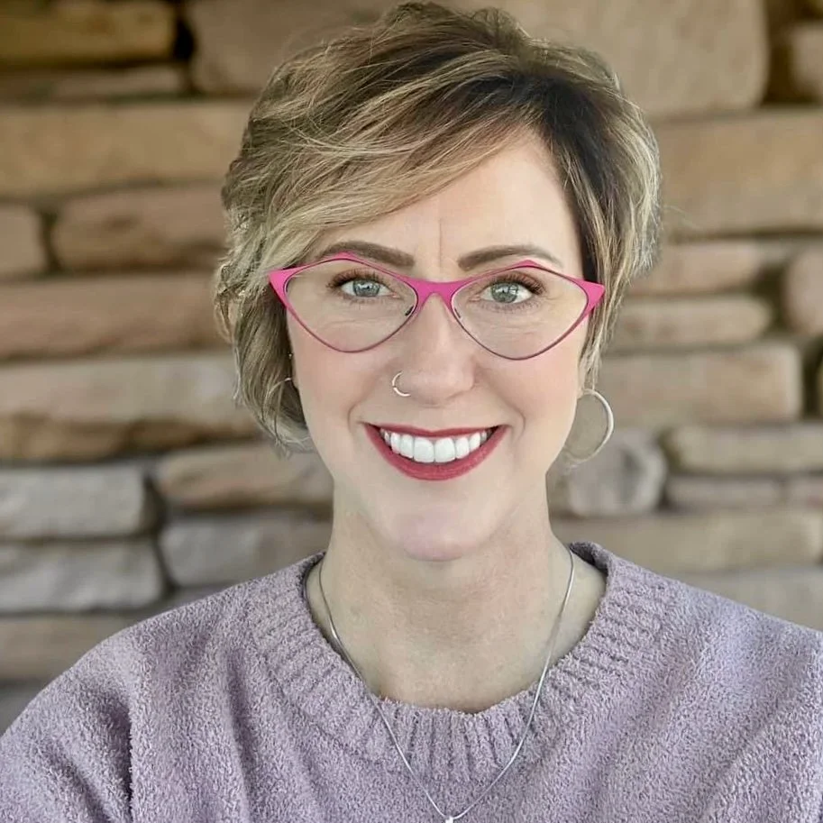A smiling woman with short, blonde hair wearing pink glasses, a nose ring, earrings, a lavender sweater, and a necklace, standing in front of a wooden wall.