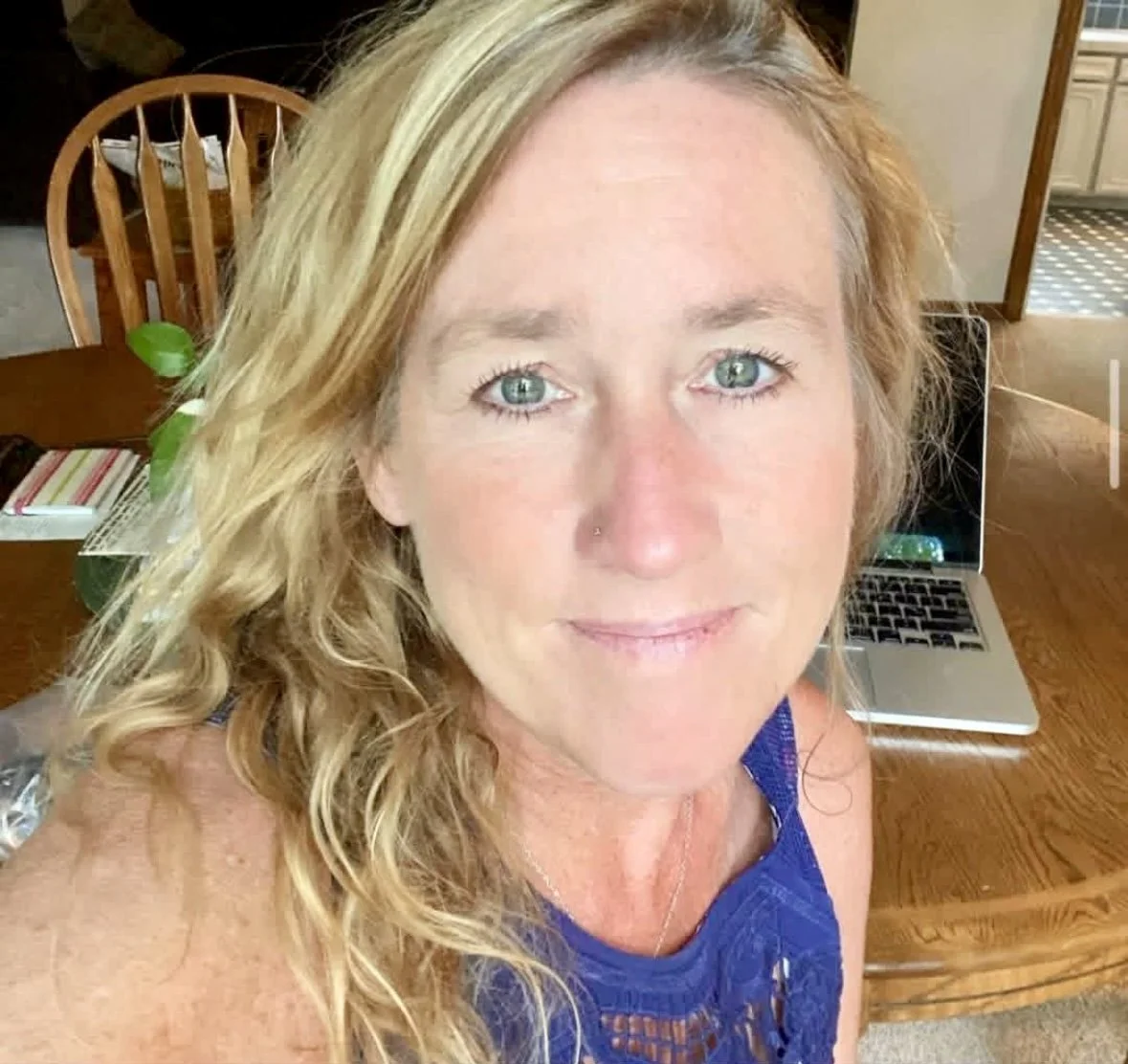 A woman with blond hair and blue eyes smiling at the camera, sitting at a wooden table with a laptop and papers in a kitchen.