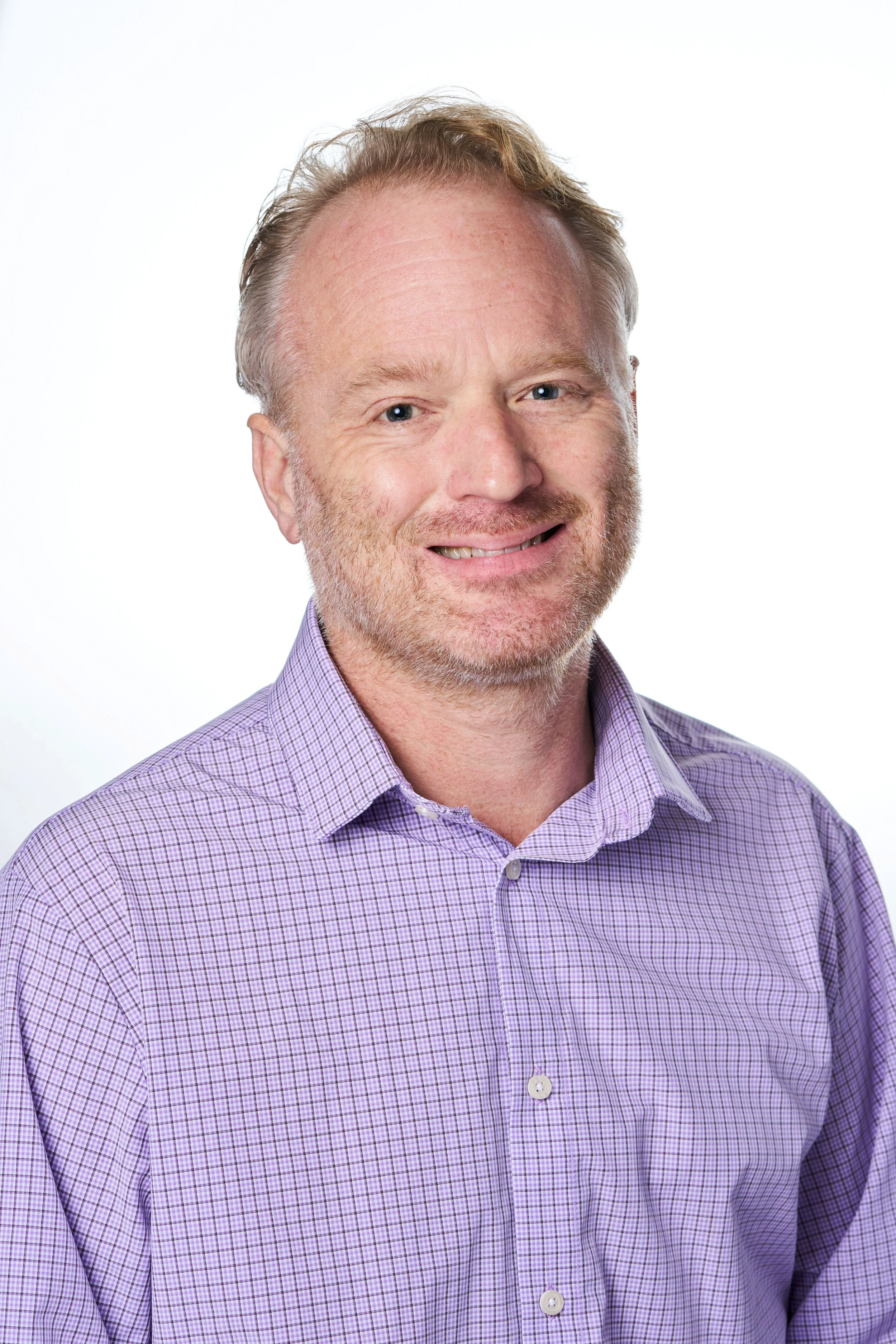 A middle-aged man with light skin, red hair, and a beard, smiling, wearing a purple checkered shirt, against a white background.