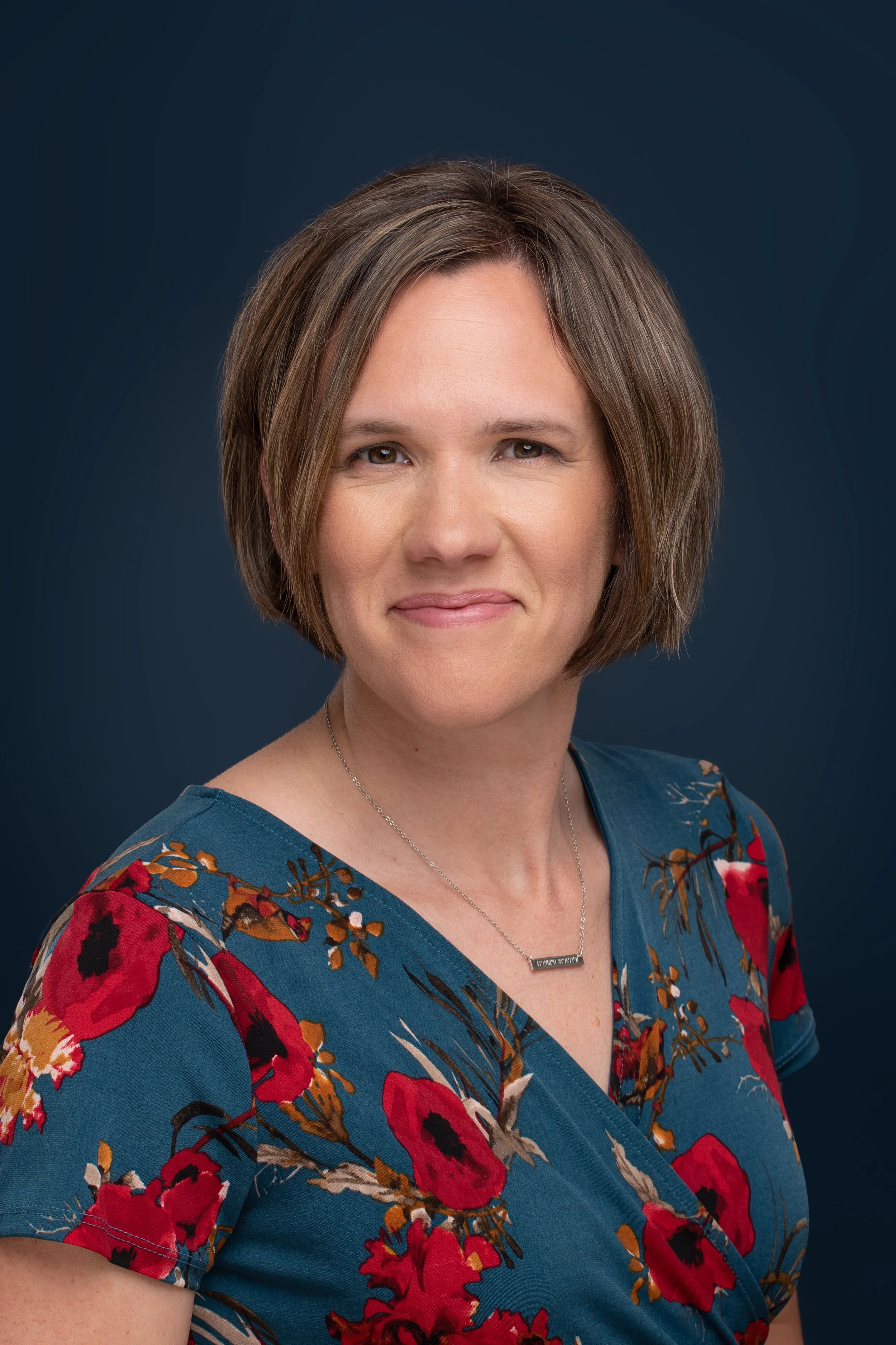 A woman with short brown hair wearing a blue floral dress and a silver necklace against a dark blue background.