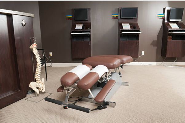 A chiropractic examination room with an adjustable treatment table, a model of a human spine on a stand, and three computer workstations along the wall.