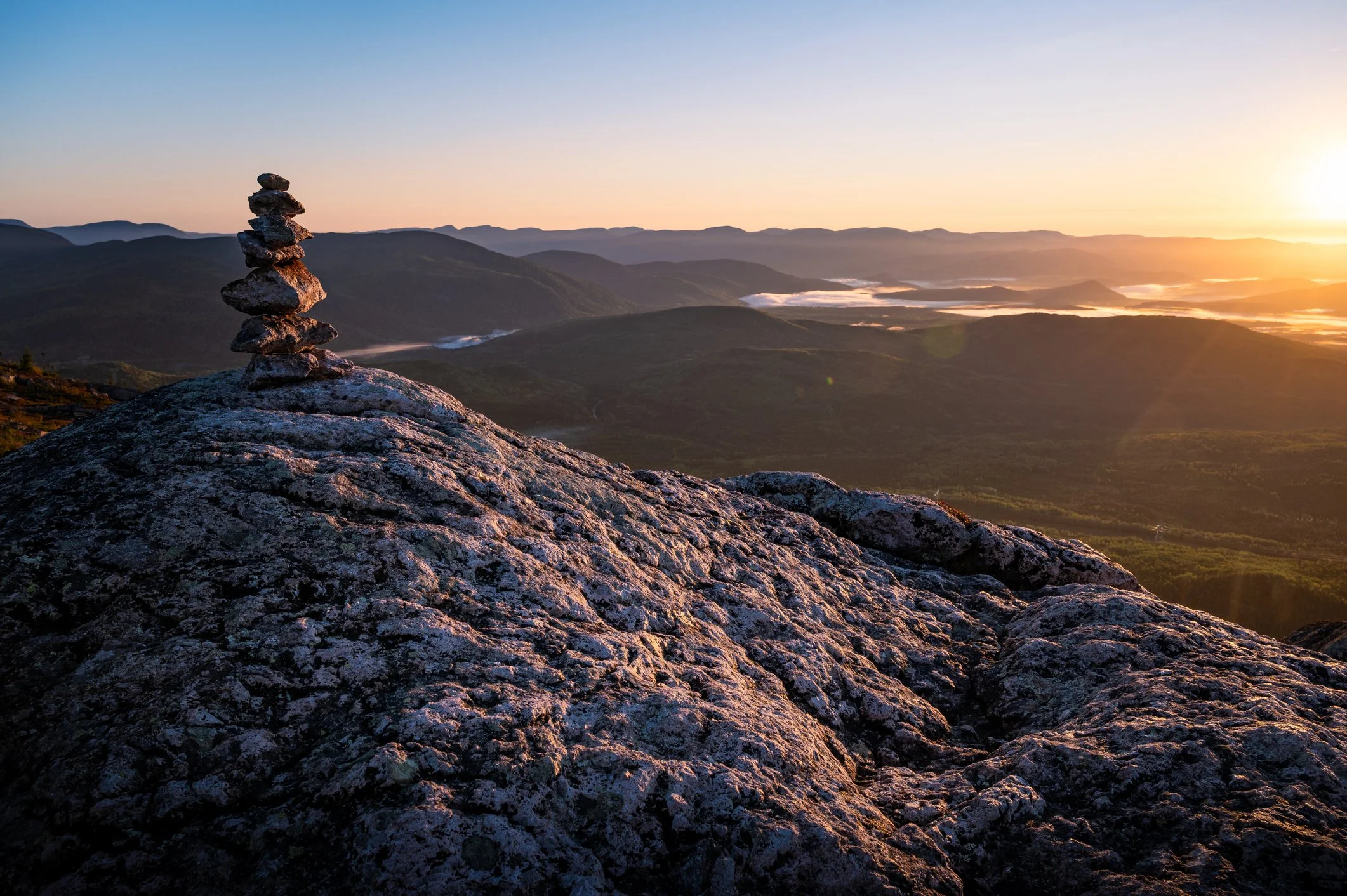 A scenic mountain landscape during sunset with a stack of balanced rocks on a large rocky surface in the foreground. Distant hills and valleys are visible under a sky with a setting sun.