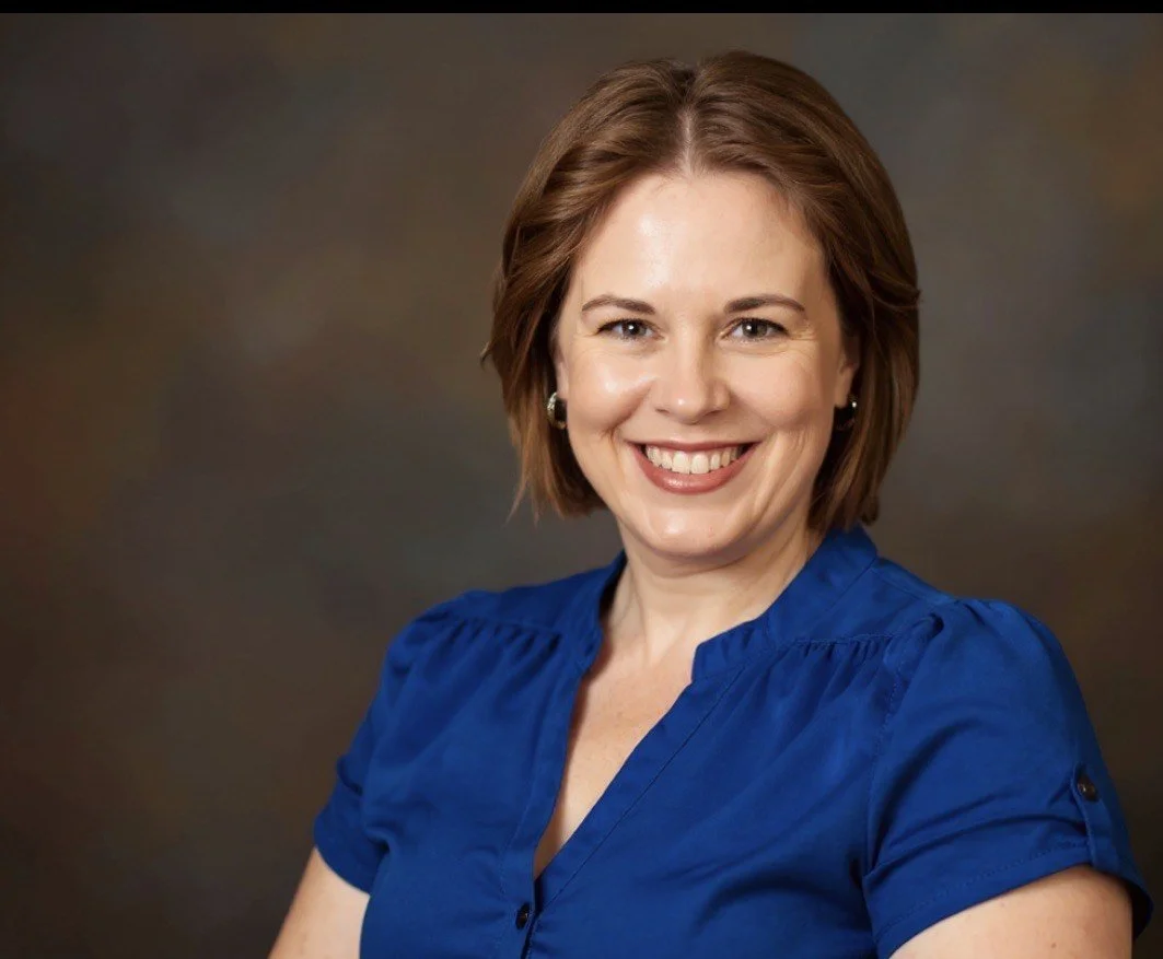 A woman with short brown hair smiling, wearing a blue blouse, against a brown background.