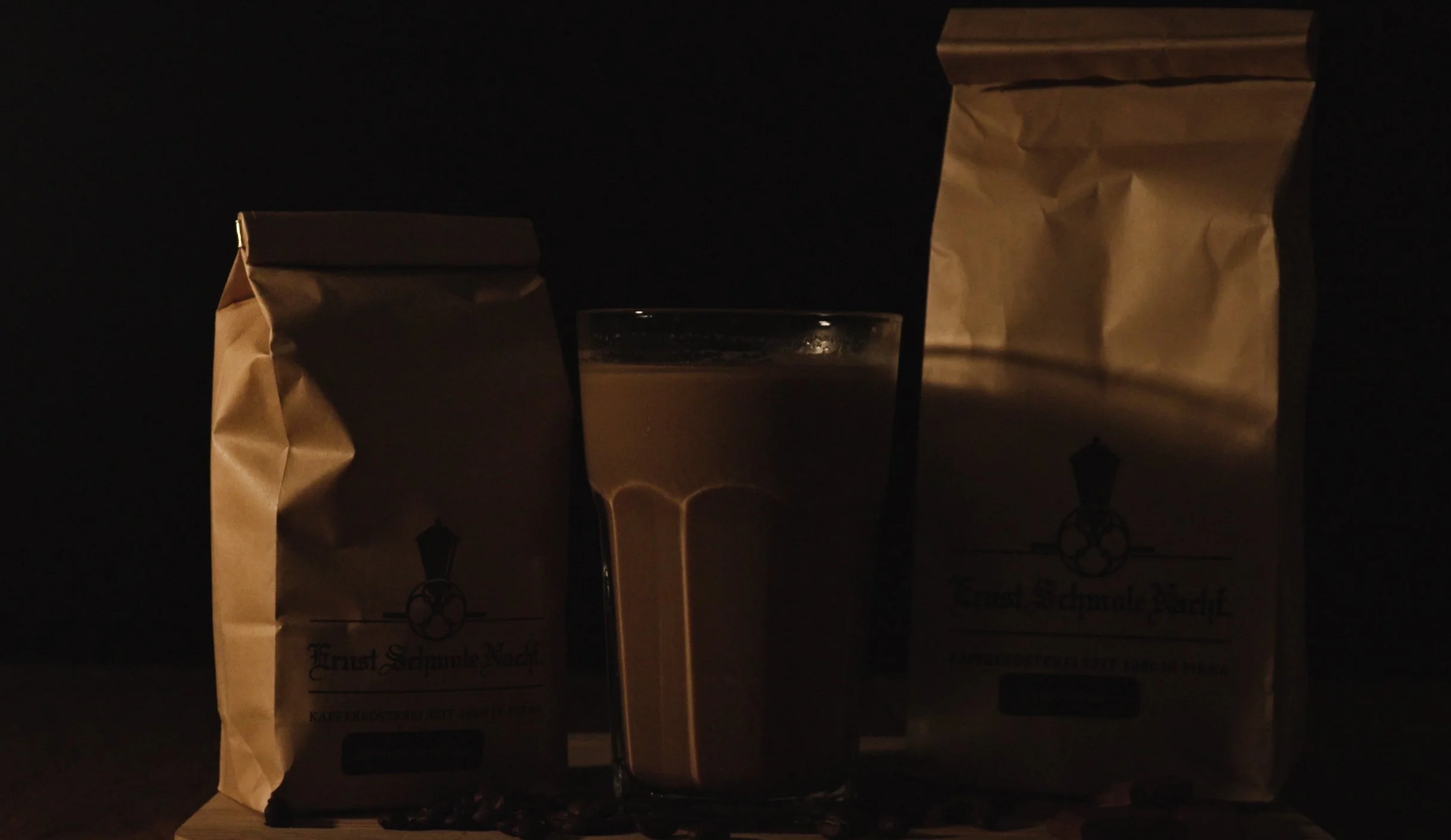 Ein Glas Kaffee mit Milch zwischen zwei Kaffeepackungen auf einem dunklen Hintergrund.