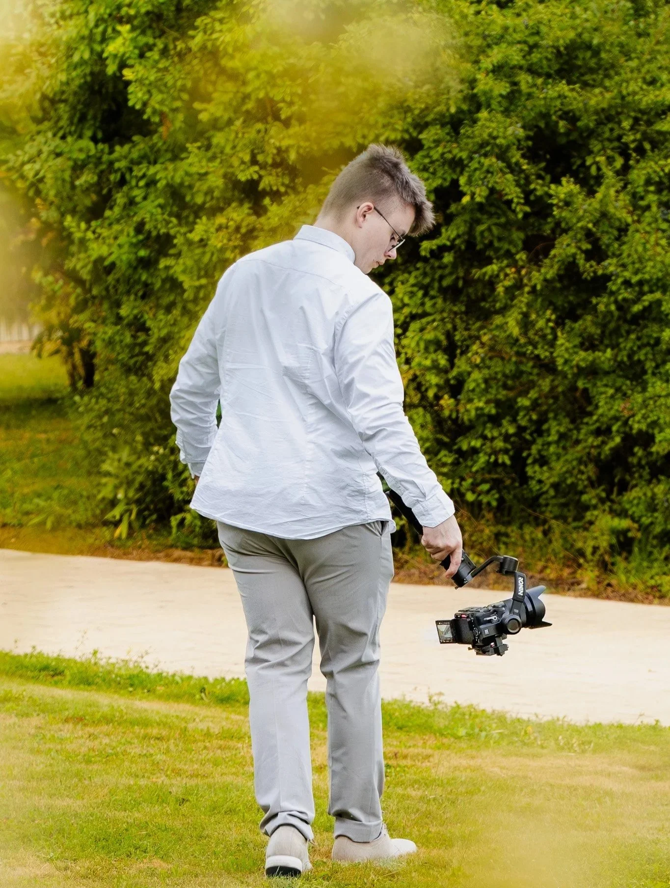 Junger Mann in weißen Hemd und grauen Hosen, der eine Kamera im Freien aufnimmt. Im Hintergrund sind Bäume und ein grüner Rasen zu sehen.