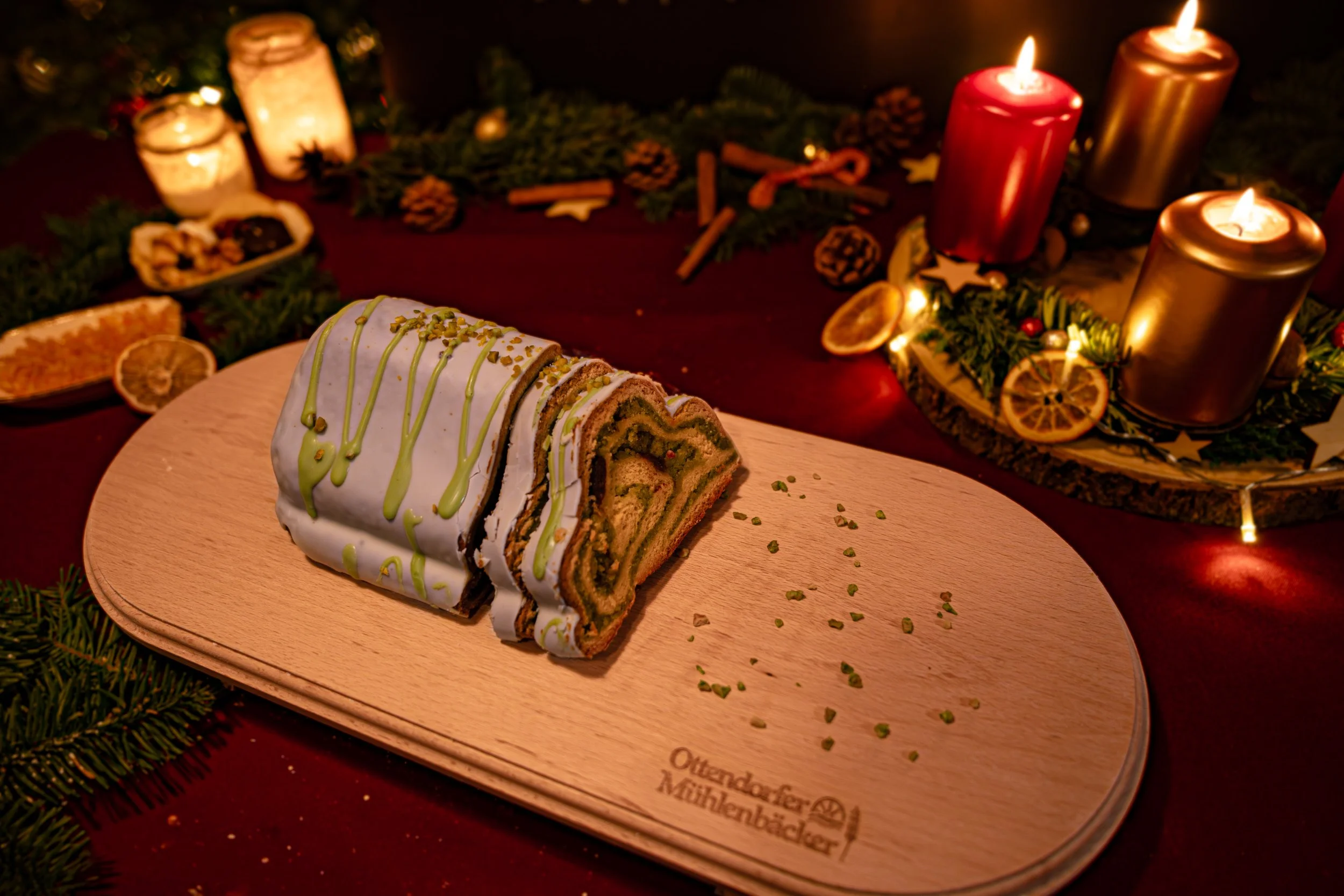 Ein Stück Weihnachtsstollen auf einem Holzbrett, umgeben von festlicher Dekoration mit Kerzen, Tannenzweigen, getrockneten Orangenscheiben und Gewürzen.