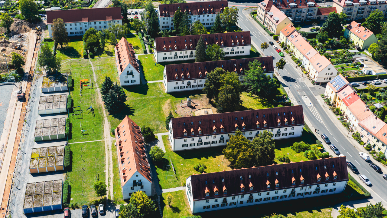 Eine Vogelperspektive auf eine Wohnanlage mit mehreren weißen Gebäuden mit roten Dächern, einem Spielplatz, Gärten und Parkplätzen, umgeben von Straßen und weiteren Häusern.