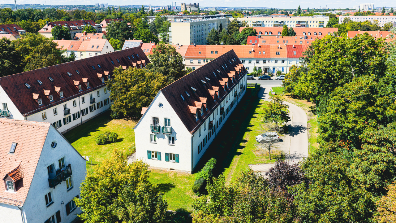 Ansicht eines Wohnviertels mit mehreren Gebäuden, gepflegten Grünflächen und Bäumen. Im Vordergrund steht ein langes, weißes Mehrfamilienhaus mit rotbraunem Dach. Im Hintergrund sind weitere Wohnblocks und ein Horizont mit Stadtgebäuden sichtbar.