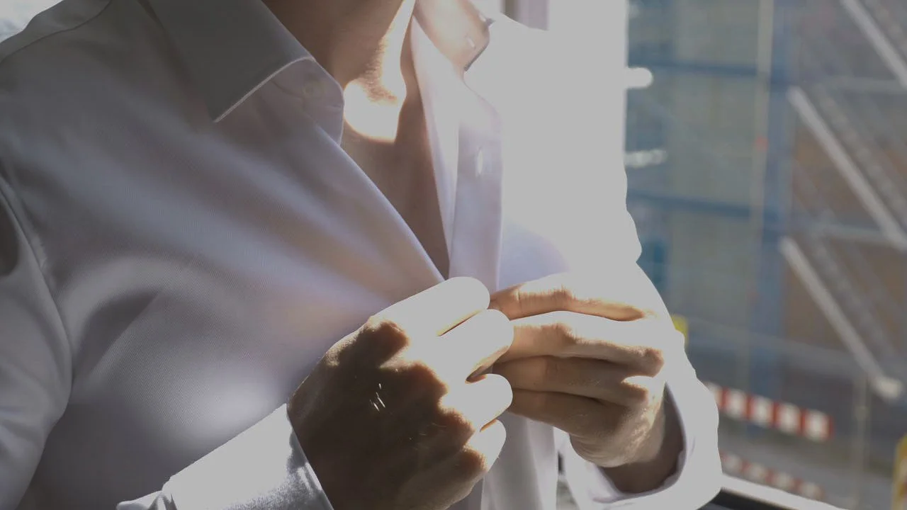 Nahaufnahme eines Mannes in weißem Hemd, der an seinem Mantel zieht, mit einem modernen Stadtbild im Hintergrund.