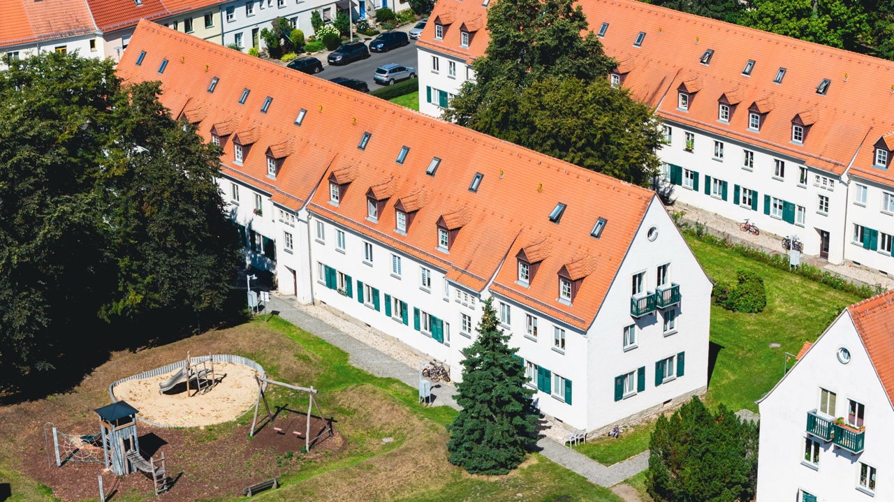 Mehrstöckige weiße Wohnhäuser mit roten Dächern, umgeben von Bäumen und einem Spielplatz im Vordergrund