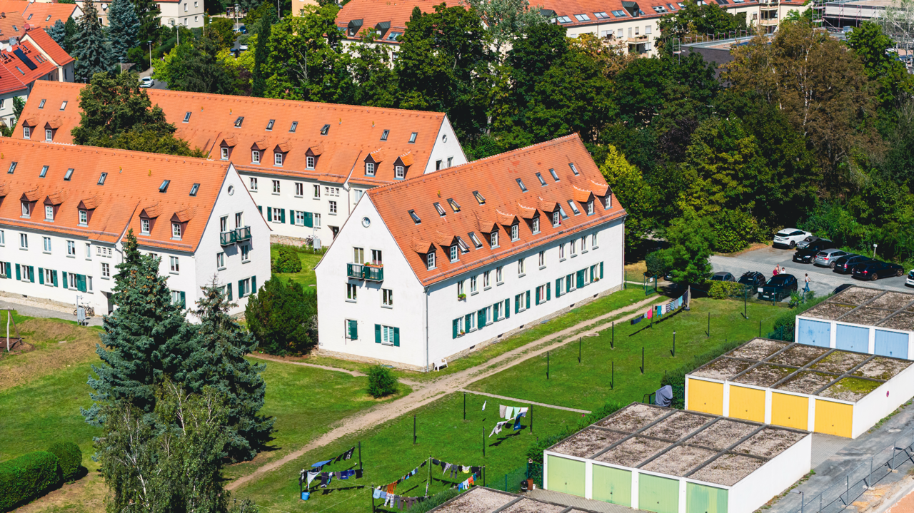 Mehrfamilienhaus mit roten Dächern und grünen Fensterläden, umgeben von Bäumen und Wiesen, mit Wäscheleinen im Garten, in einer urbanen Wohngegend.