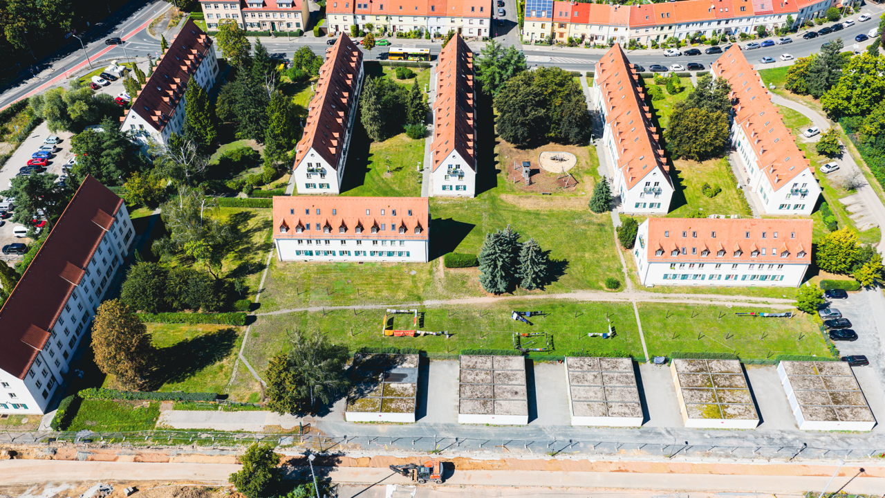 Luftaufnahme eines Wohnviertels mit mehreren weißen Mehrfamilienhäusern mit roten Dächern, Spielplätzen im grünen Garten und parkenden Autos auf den Straßen.