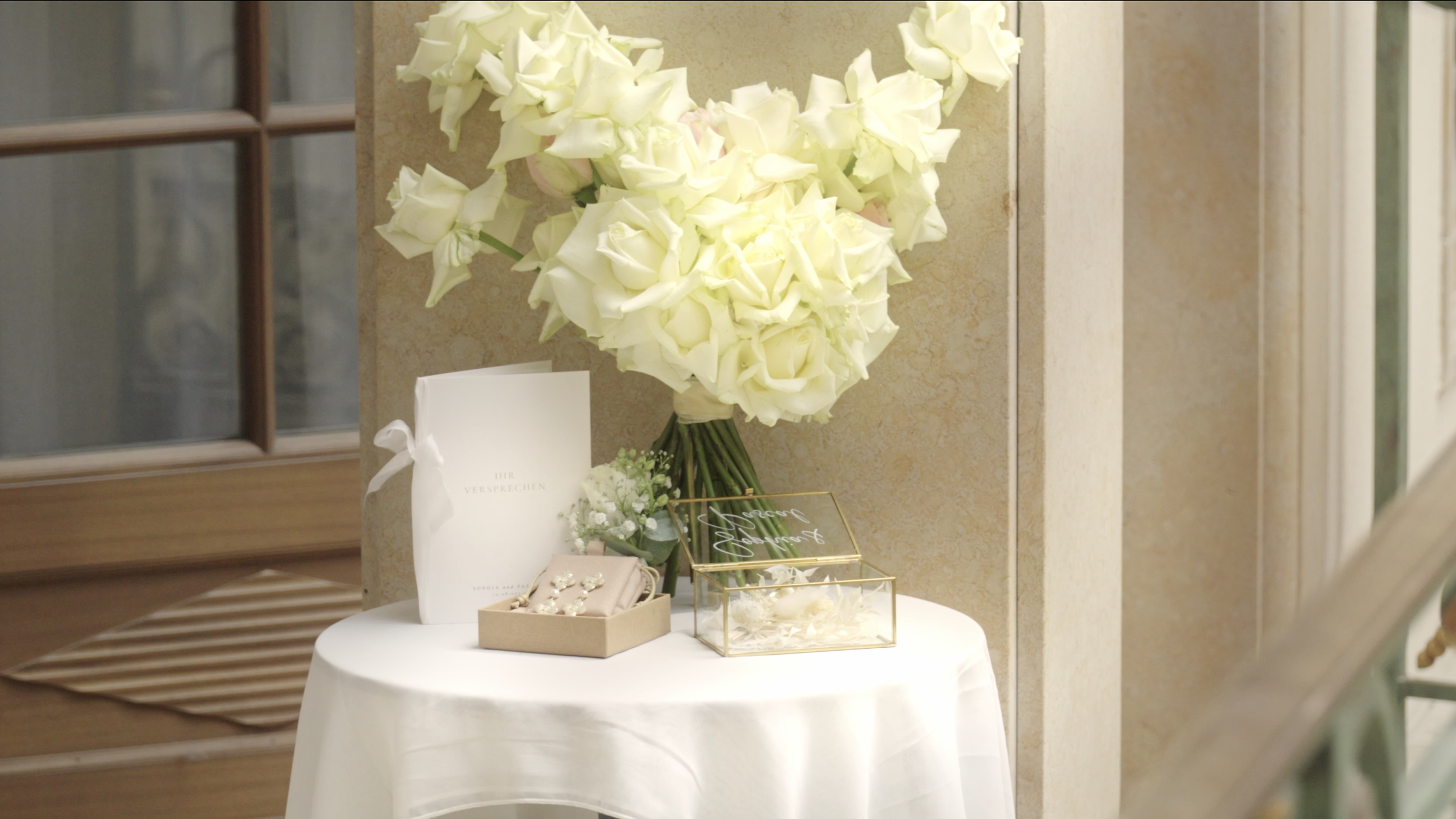 Ein Blumenstrauß aus weißen und zartrosa Rosen auf einem weißen Tisch. Daneben liegen Geschenkverpackungen und eine kleine Schachtel mit Schmuck. Im Hintergrund eine Tür mit Glasfenster