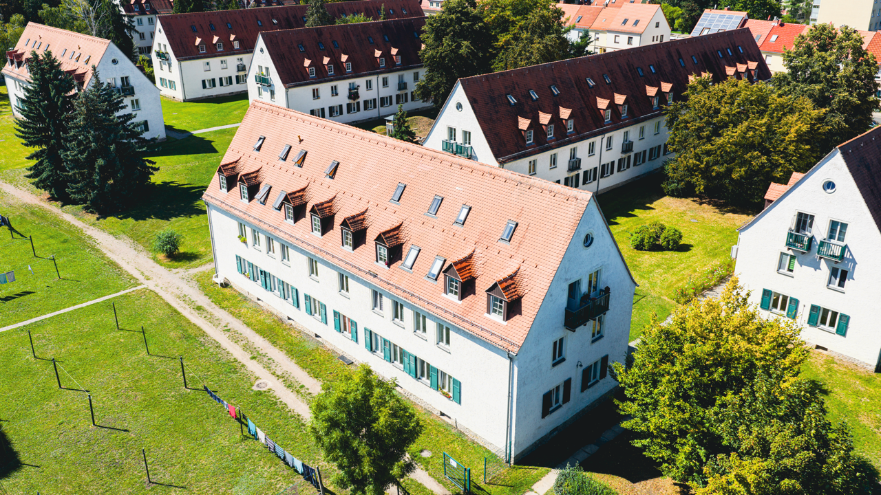 Luftaufnahme einer Wohngegend mit weißen Mehrfamilienhäusern, roten Dachziegeln, grünen Rasenflächen und Bäumen.
