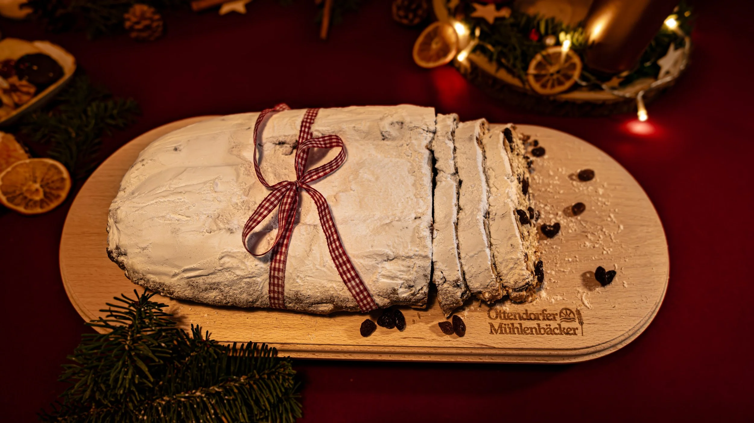 Geschnittenes Weihnachtsgebäck auf einem Holzbrett, dekoriert mit einer roten Schleife, umgeben von Weihnachtsdeko und Tannenzweigen, mit Sebstbezeichnung "Ottendorfer Mühlenbäcker".