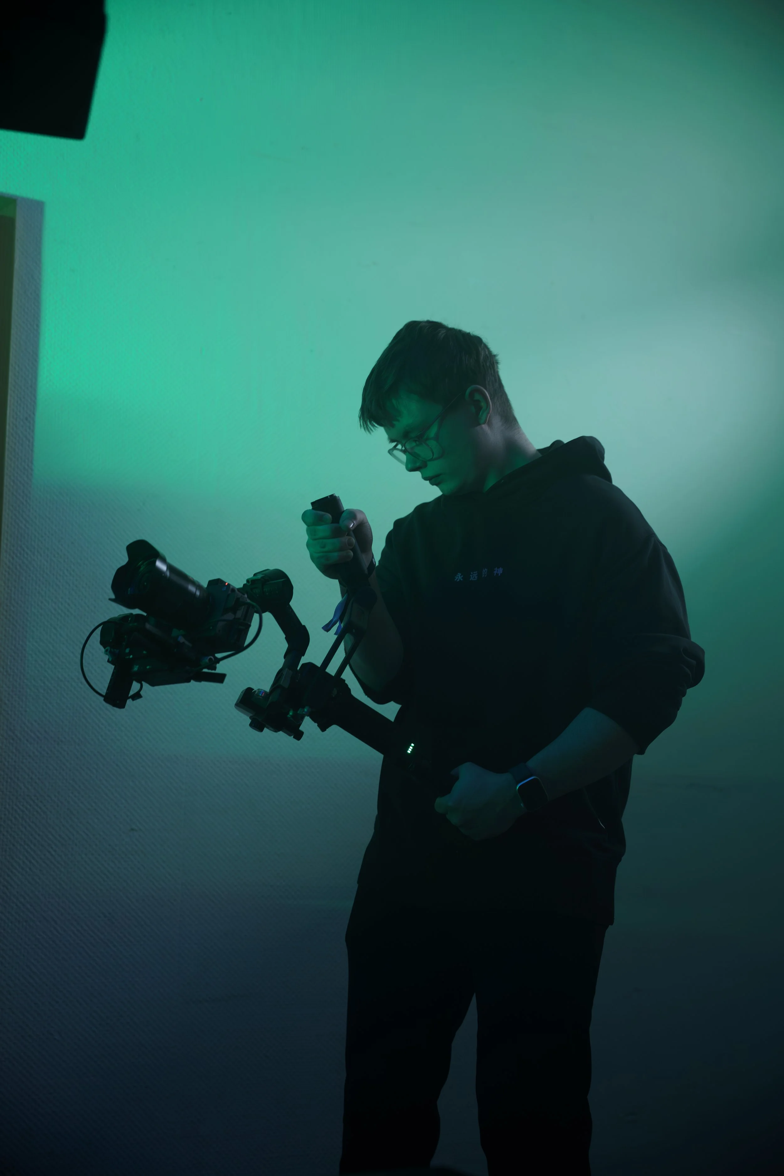Ein junger Mann mit Brille hält eine Kamera mit Gimbal in einer dunkelgrauen Raum, bei dem grünes Licht an ist.