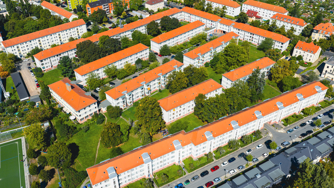 Luftaufnahme einer Wohnsiedlung mit mehreren weißen Gebäuden mit roten Dächern, umgeben von Bäumen und Straßen mit parkenden Autos.