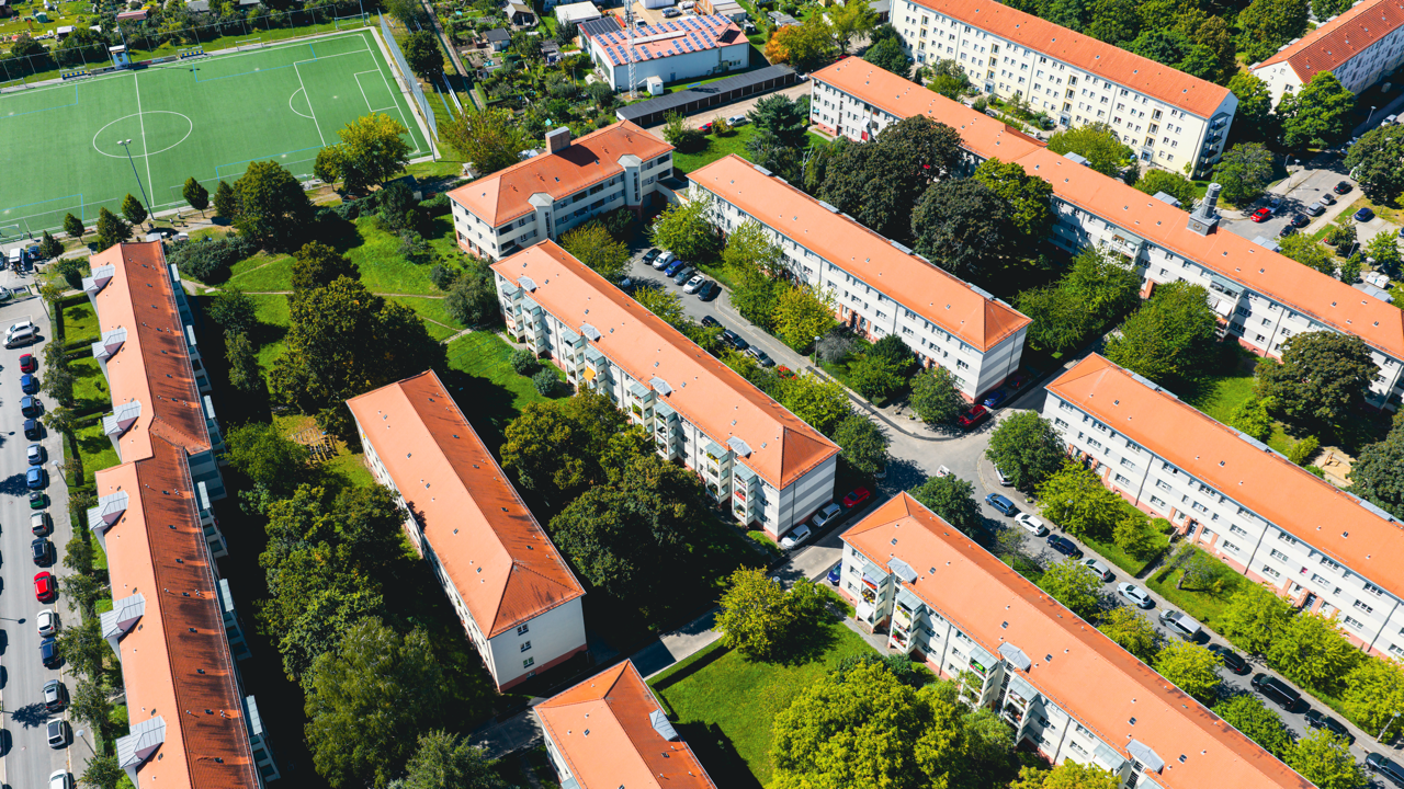 Luftaufnahme eines Wohnviertels mit mehreren Mehrfamilienhäusern mit roten Dächern, umgeben von Bäumen und Straßen. Im oberen linken Bereich befindet sich ein Fußballfeld.
