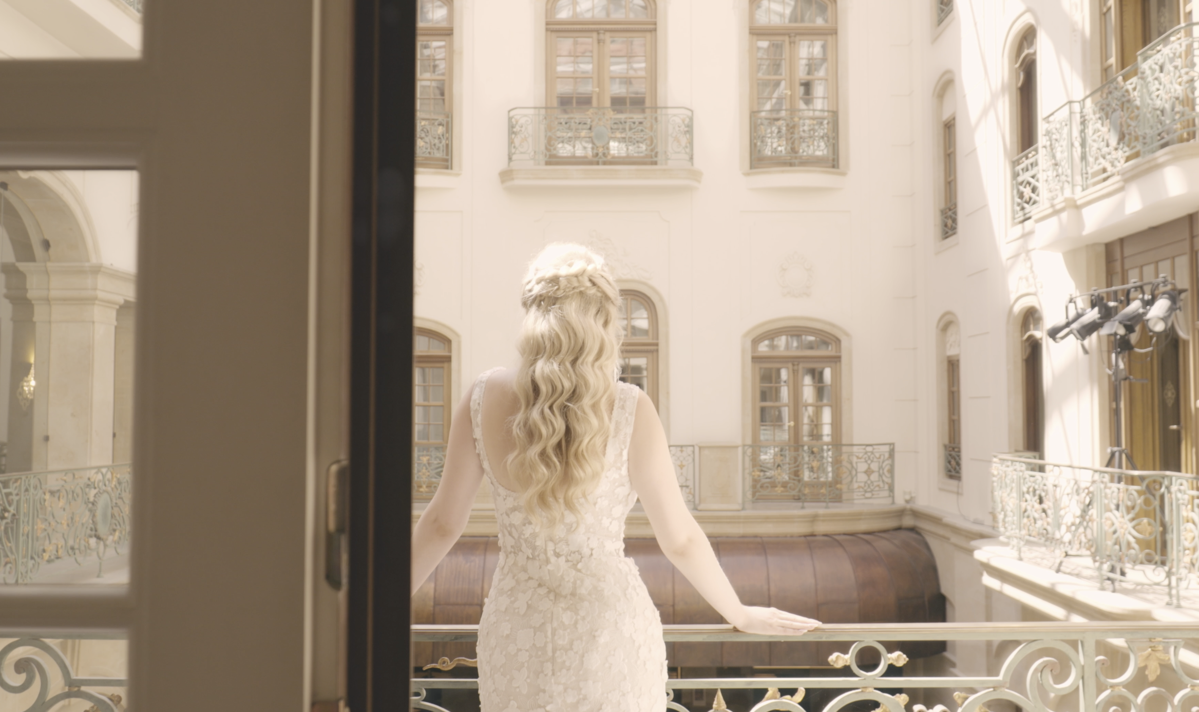 Frau in Brautkleid blickt aus einem Balkon einer eleganten, hell erleuchteten, historischen Innenarchitektur mit mehreren Fenstern und Balkonen.