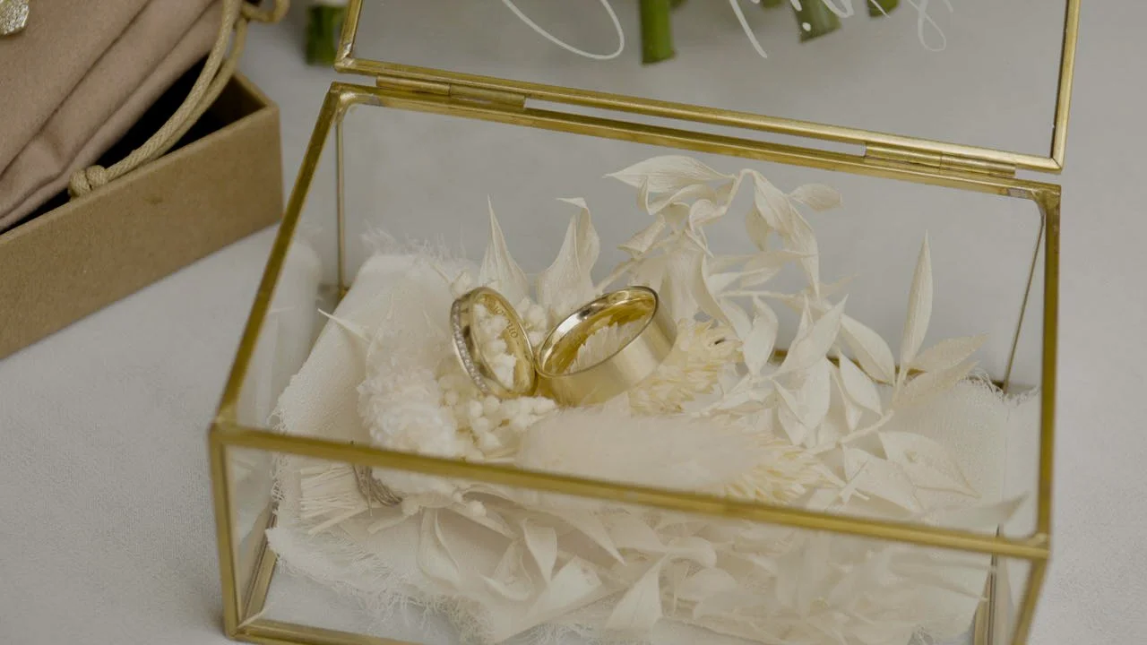 Zwei goldene Eheringe in einem goldfarbenen Schmuckkästchen auf weißem Dekor, umgeben von weißen Blättern.