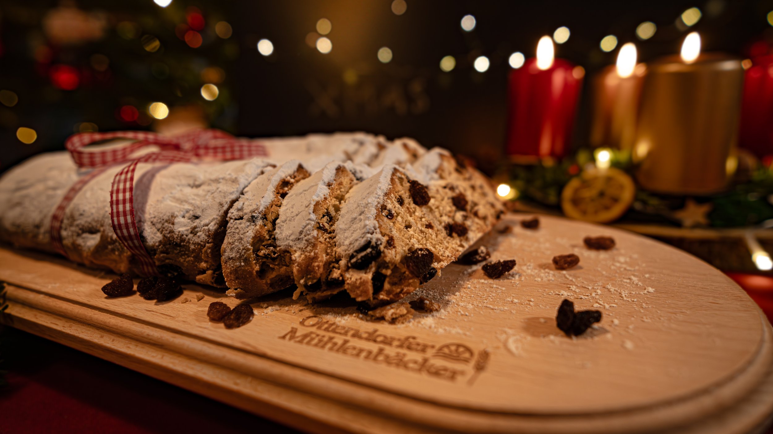 Ein geschnittener Christstollen mit Puderzucker auf einem Holzbrett, im Hintergrund Weihnachtsdeko und Kerzen.