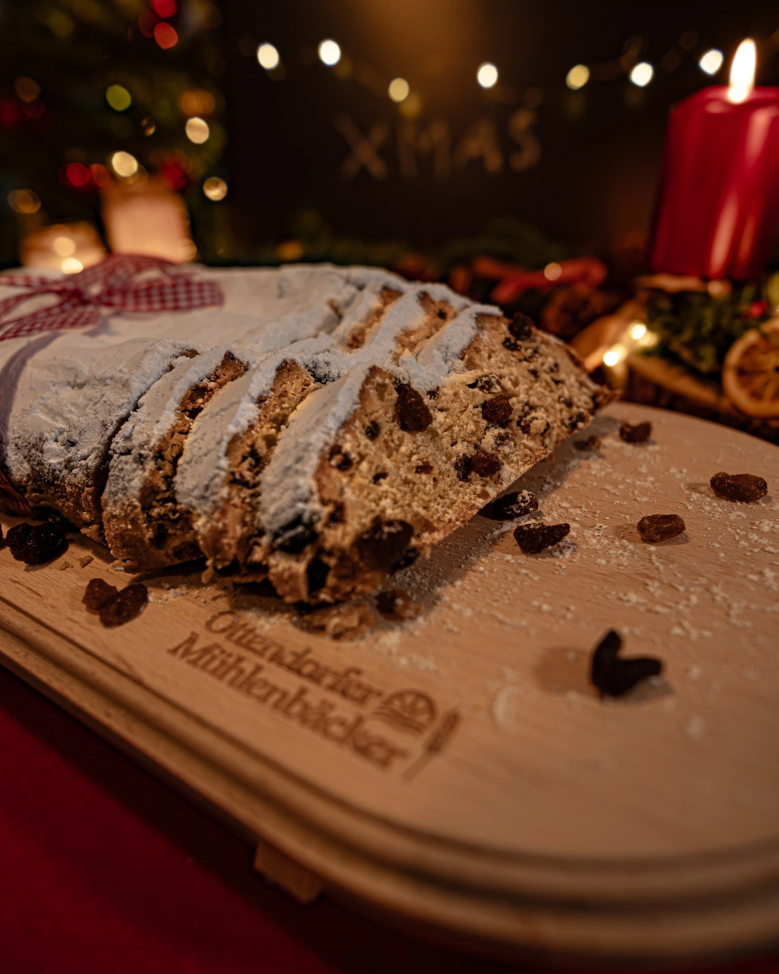 Weihnachtsstollen auf Holzbrett, mit Puderzucker bestäubt, in einer festlichen Weihnachtsdekoration im Hintergrund, inklusive Kerzen und Geschenken.