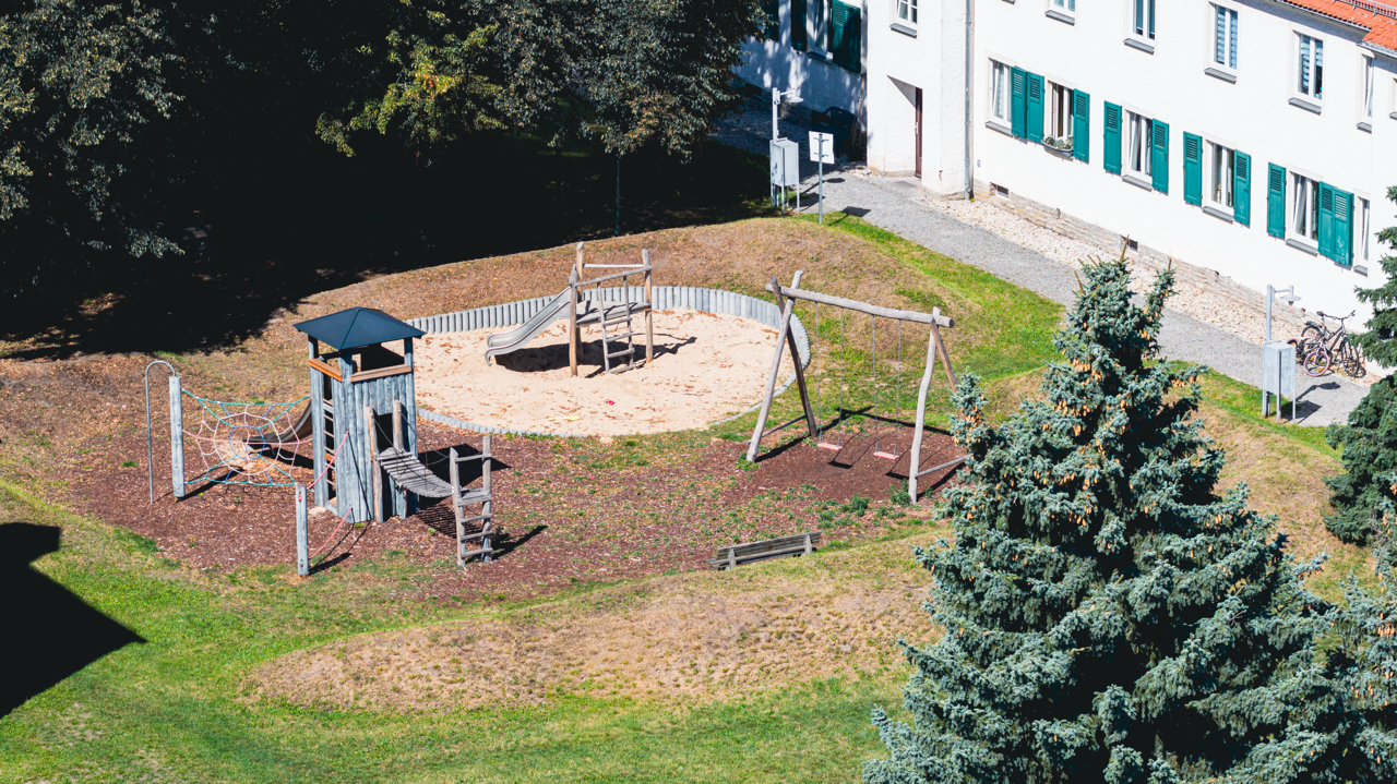 Hübscher Spielplatz mit Sandkasten, Rutsche und Schaukeln in einem Grünbereich neben einem weißen Mehrfamilienhaus mit grünen Fensterläden.