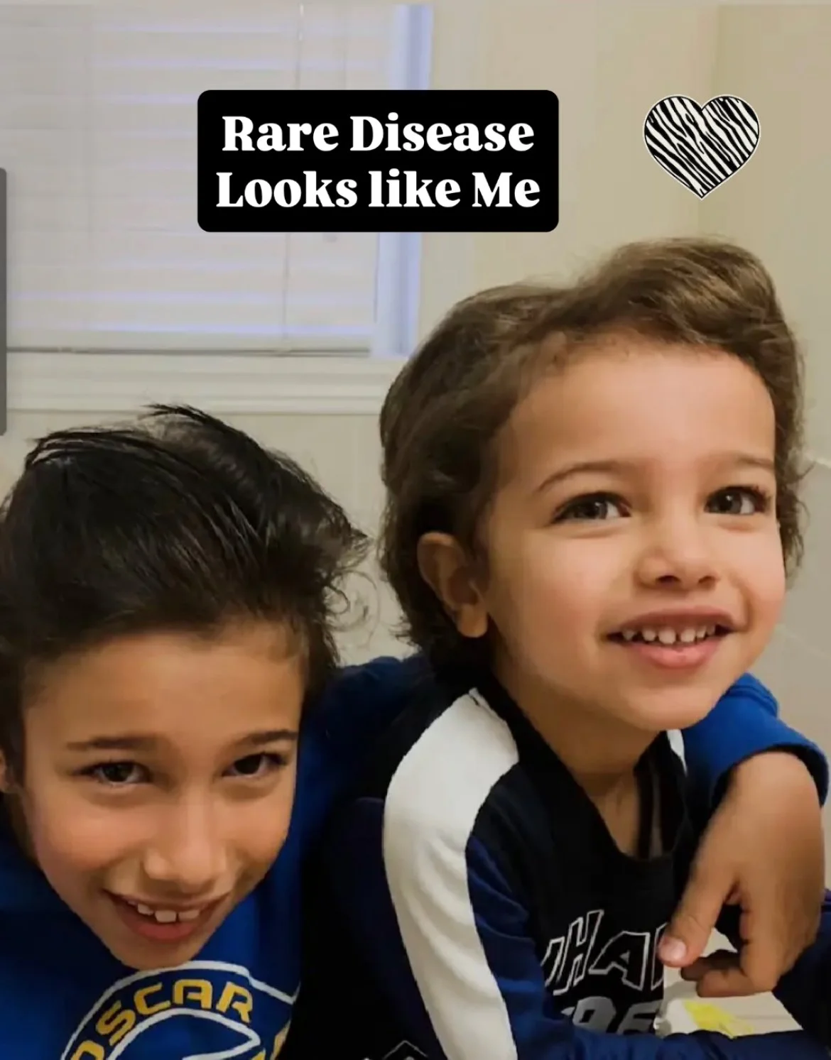 Two smiling kids, one with dark hair, wearing a blue hoodie with a logo, and the other with light brown hair, wearing a black and white long-sleeve shirt, sit close together indoors. The image has a caption saying 'Rare Disease Looks like Me' with a black heart graphic.
