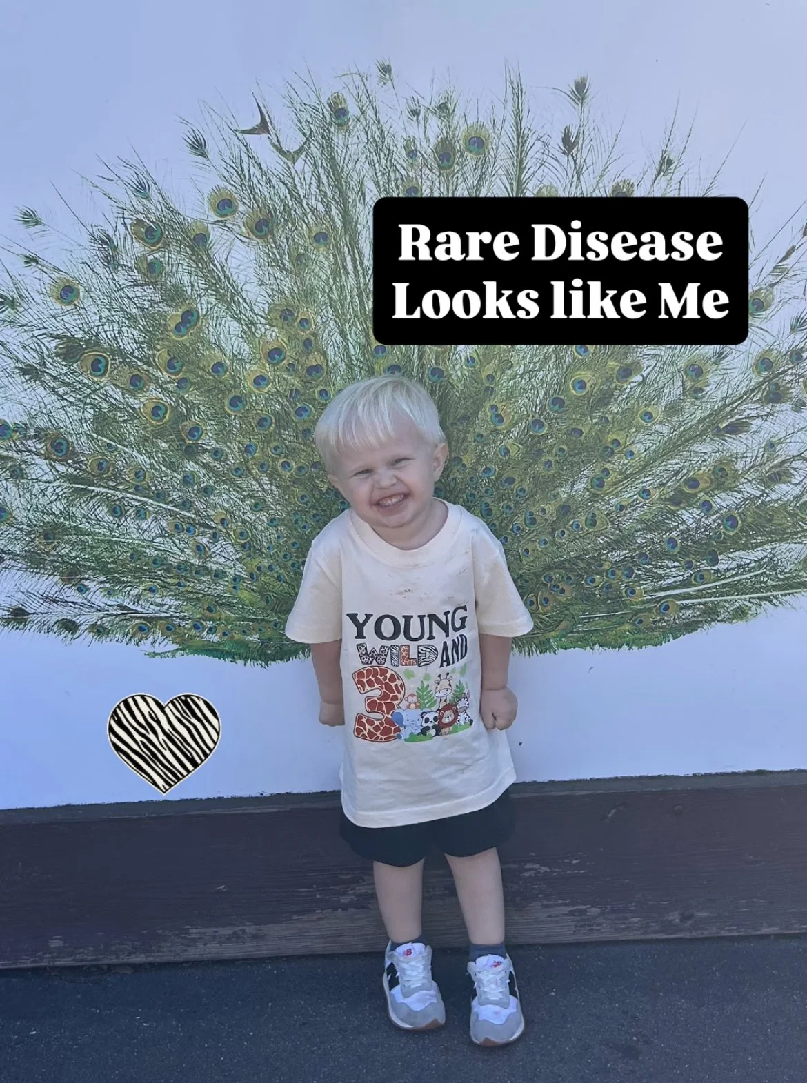 A young boy with blonde hair smiling and squinting, standing in front of a peacock mural that has its tail fanned out. The boy is wearing a white T-shirt with colorful text and cartoon animals, black shorts, white sneakers, and socks. There is a black and white striped heart sticker and a black box with white text saying "Rare Disease Looks Like Me".