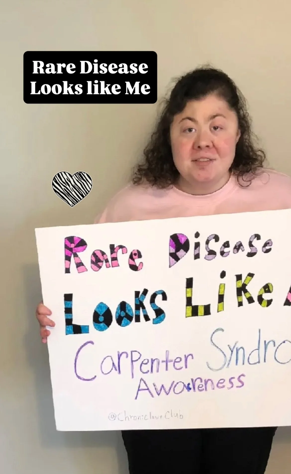 A woman holding a sign that reads 'Rare Disease Looks Like A Carpenter Syndrome Awareness', with colorful and patterned text, and a heart drawing with zebra stripes, standing against a plain wall.