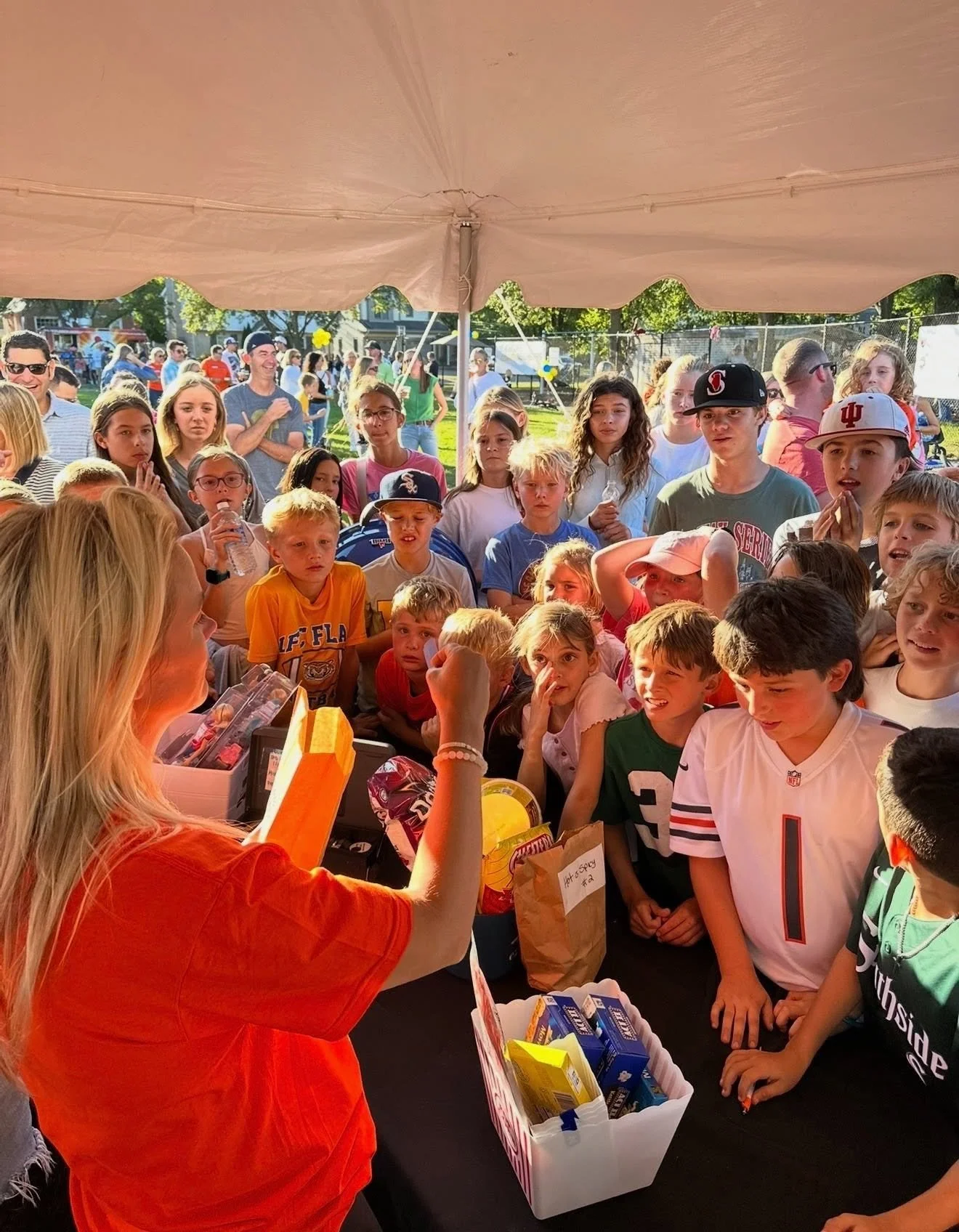 A group of children gathered around a table at an outdoor event, listening to a woman in an orange shirt who appears to be handing out snacks or supplies.
