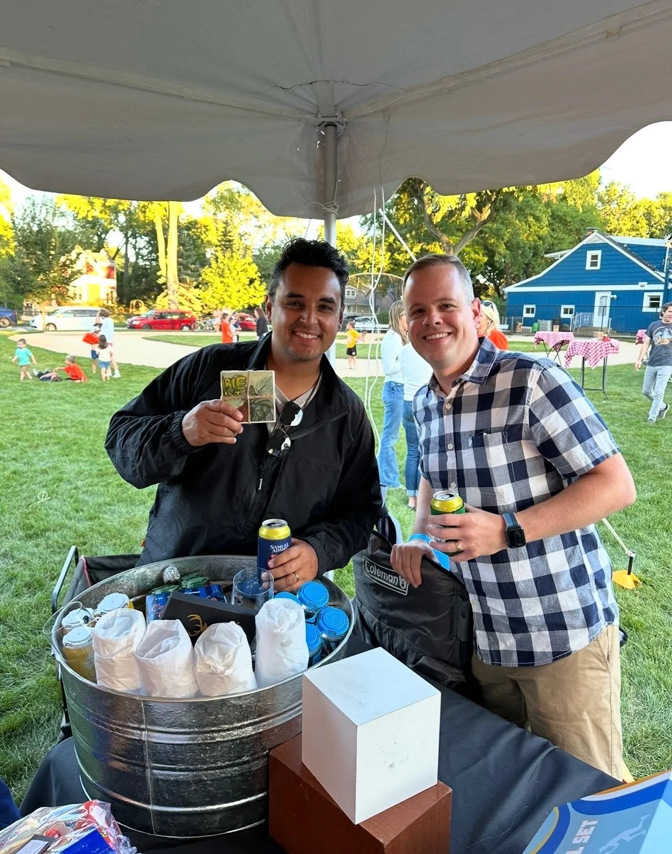 Two smiling men holding drinks at an outdoor gathering, with a large container of ice and cans, and a crowd of people in the background at a park during daylight.