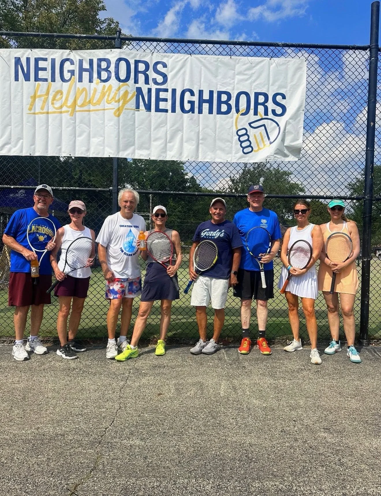 A group of nine people standing on a tennis court holding tennis rackets, smiling, with a large banner reading 'Neighbors Helping Neighbors' hanging on a fence behind them, under a partly cloudy sky.