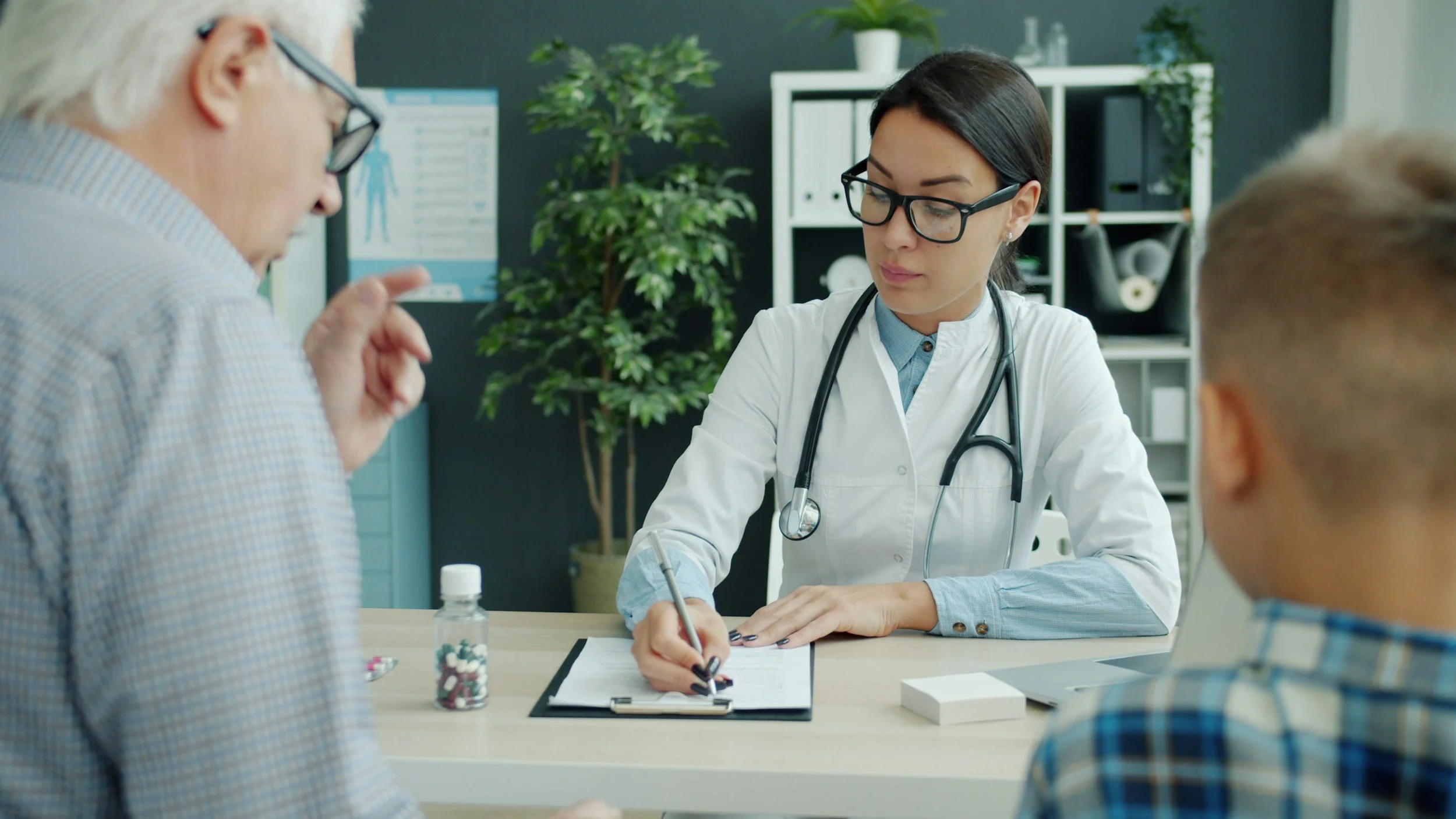 A female doctor with dark hair and glasses taking notes on a clipboard during a consultation with an elderly man and a young man in a medical office. Physicians are busy helping people. Let us help you on your financial journey.