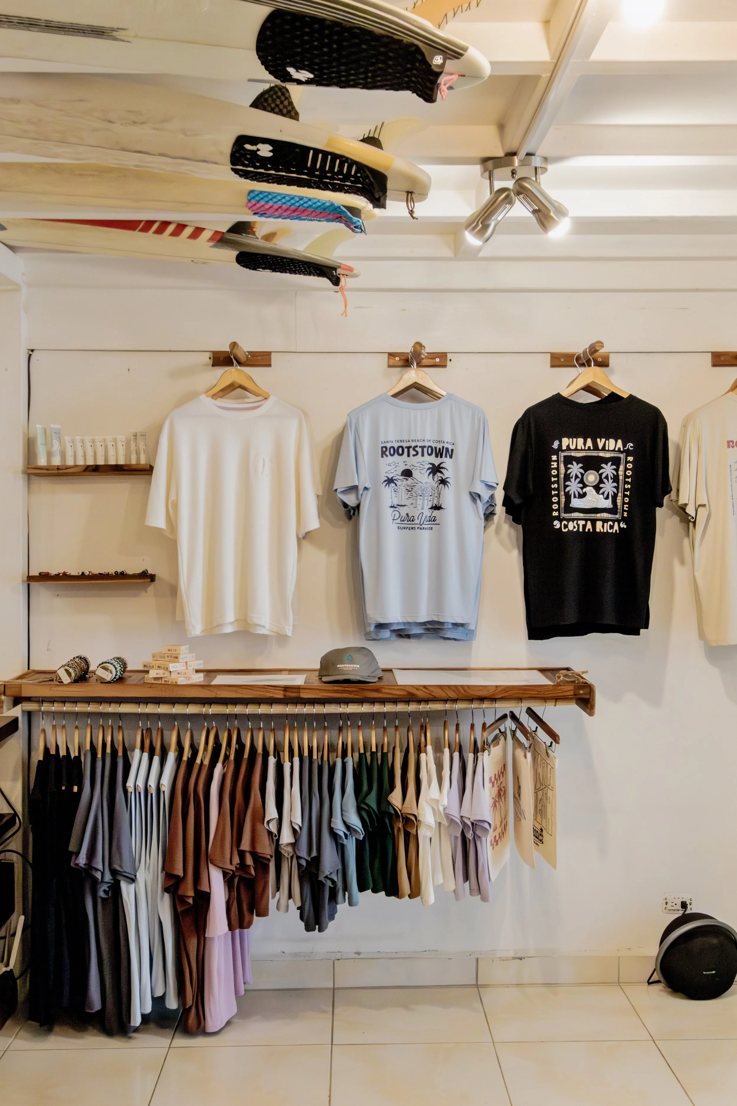 Display of surfing-related clothing and accessories, including surfboards on the ceiling, T-shirts on the wall, and shorts below, in a retail store.