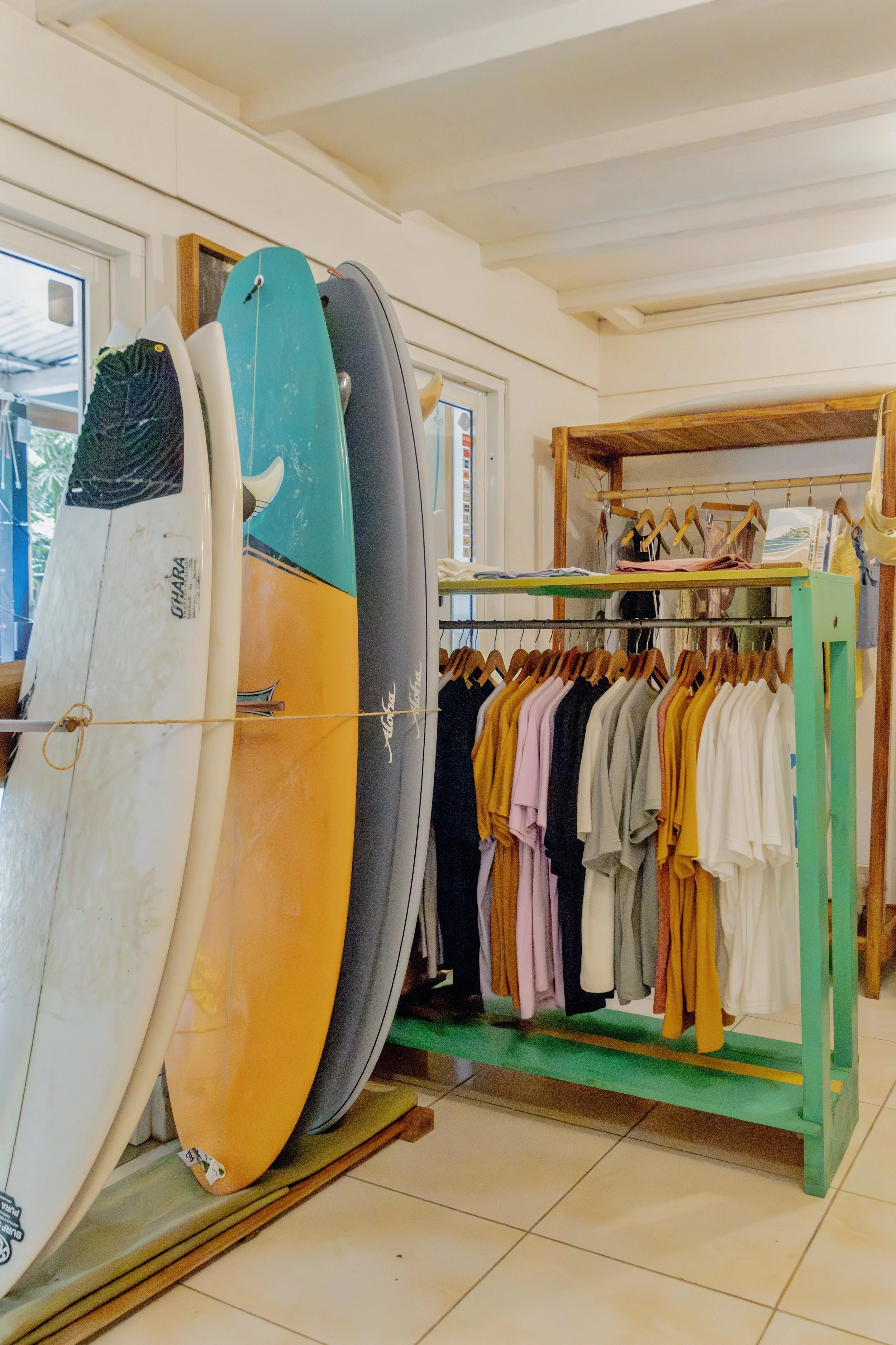 Surfboards leaning against a wall next to a clothing rack with assorted T-shirts and tops in a store or boutique.