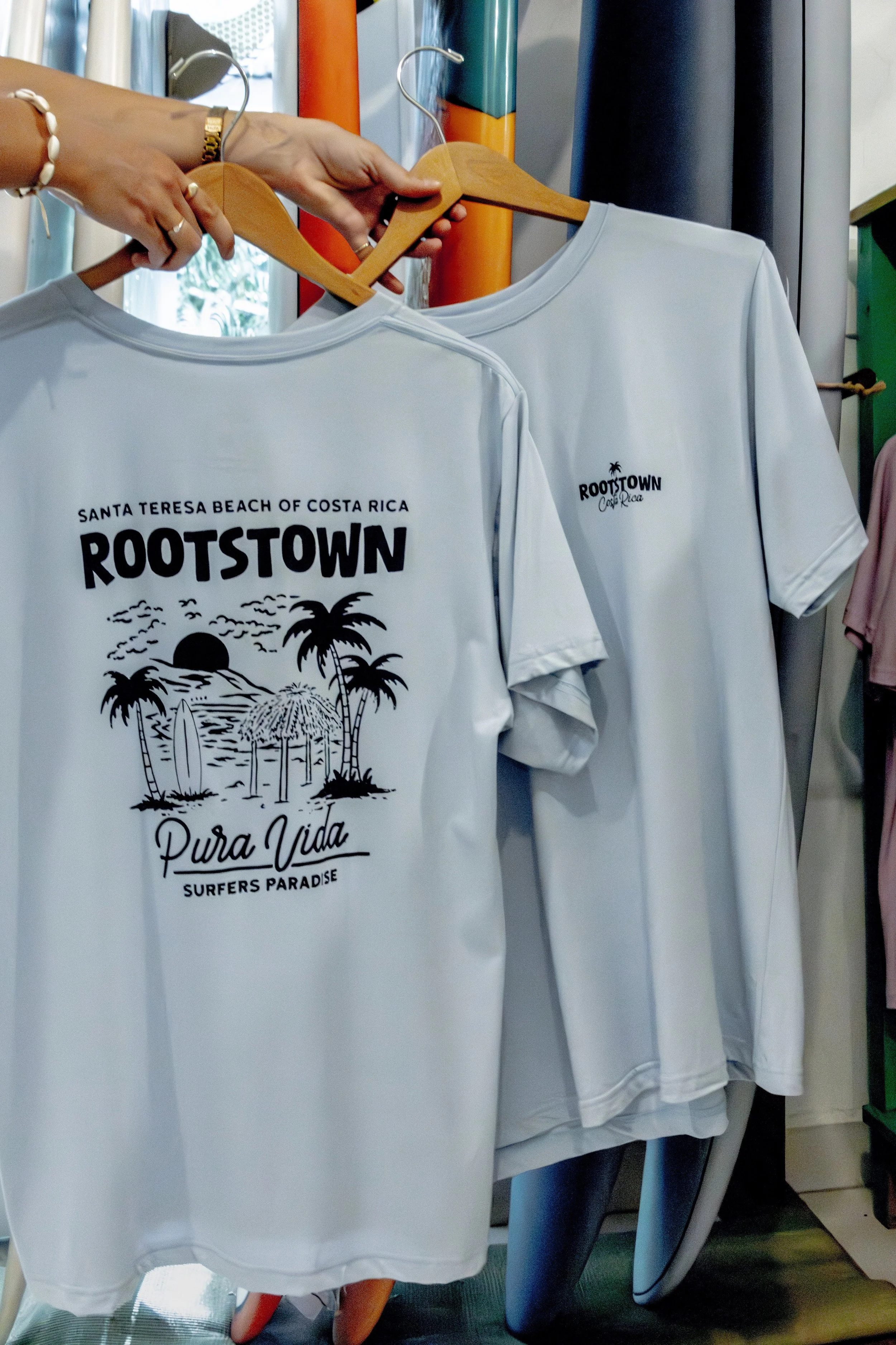 White T-shirts hanging on a rack, one with a graphic design of palm trees, a setting sun, and the words 'Santa Teresa Beach of Costa Rica, Rootstown, Pura Vida, Surfers Paradise'.