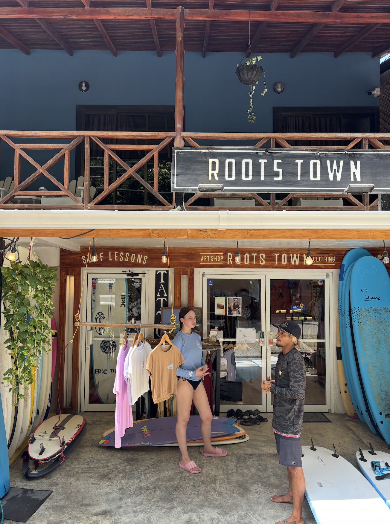 Surf shop with surfboards and apparel, people talking outside, sign reading 'ROOTSTOWN'