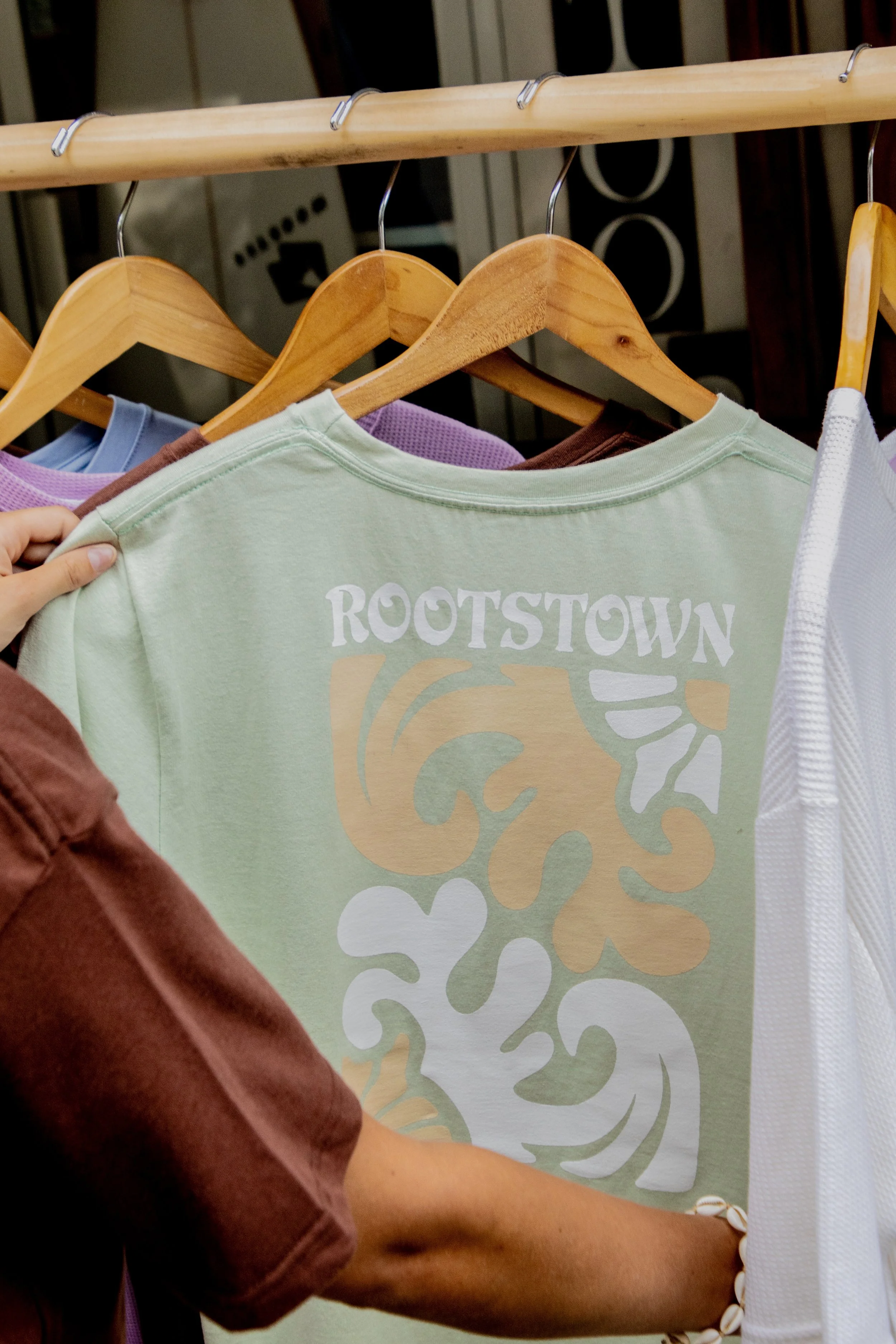 A person shopping for clothes, holding a light green T-shirt with a design that includes the word 'ROOTSTOWN' and abstract shapes, hanging on a wooden hanger among other garments.