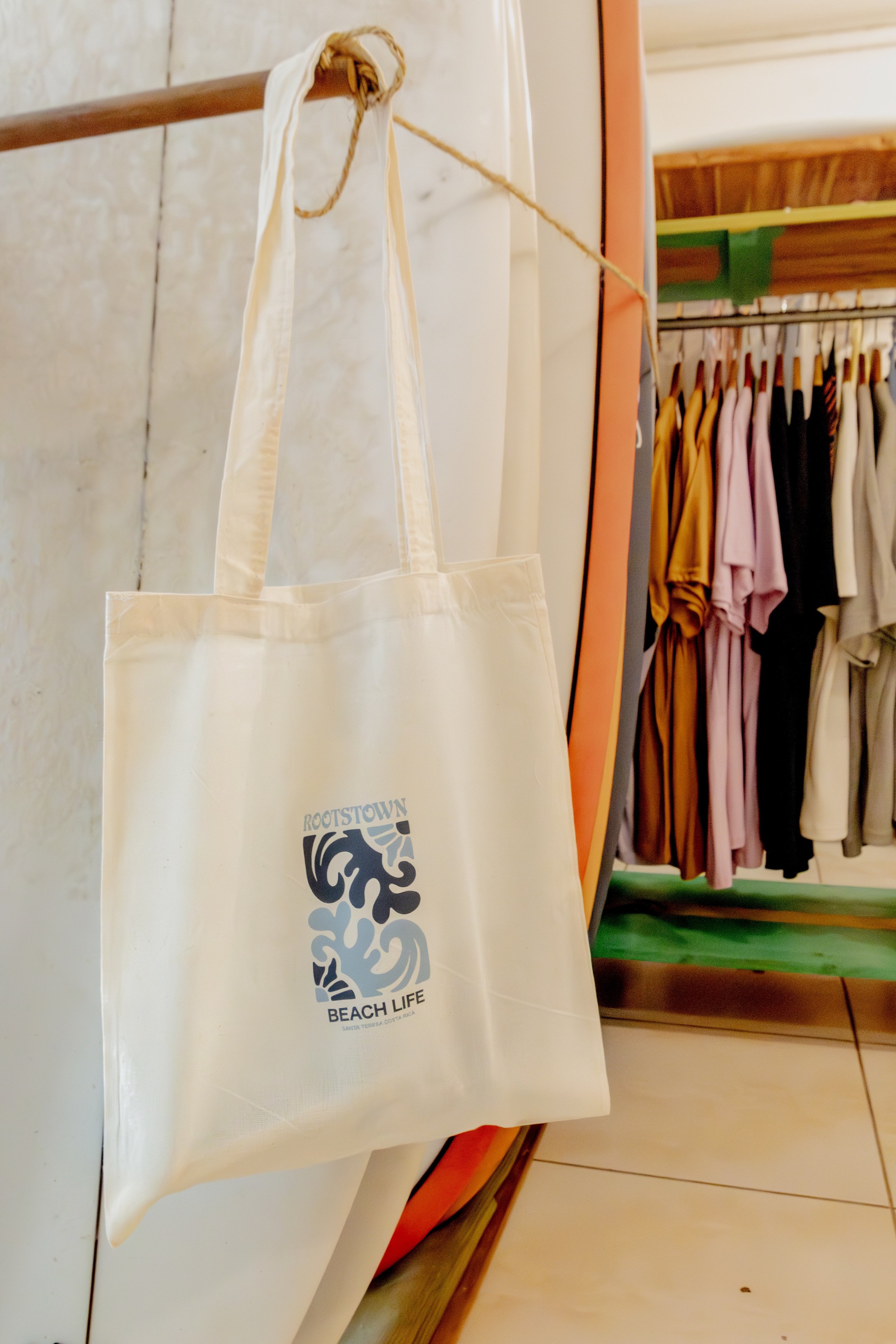 A beige tote bag hanging on a line in a clothing store with a logo that reads 'Rootstown Beach Life'. In the background, there are various colored shirts hanging on a rack.
