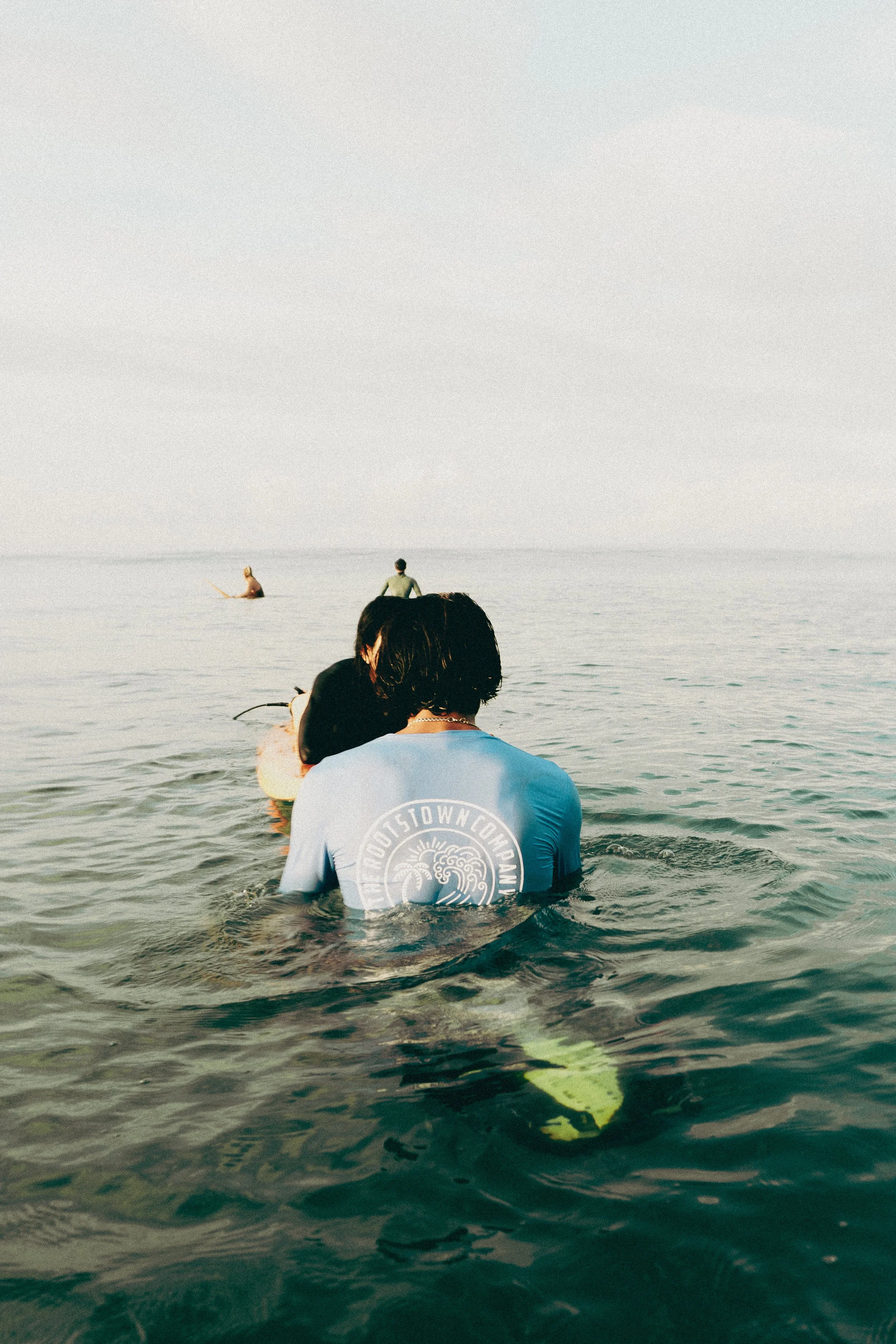 People in the ocean, with two individuals face to face in the foreground, one with wet black hair wearing a gray T-shirt and the other with dark hair, and two others in the distance paddleboarding on calm water under a cloudy sky.