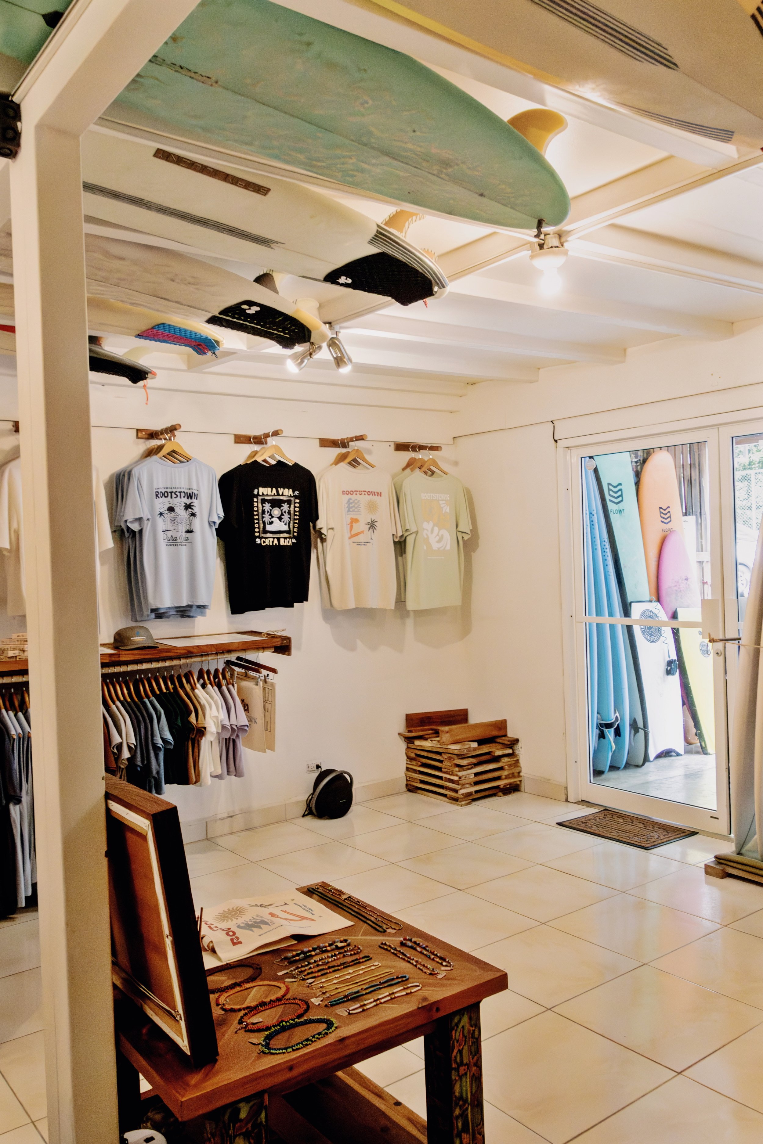 Surfboard shop interior with surfboards on the ceiling and surf-themed t-shirts hanging on the wall, displayed alongside accessories on a table and a sliding glass door leading outside, revealing more surfboards.