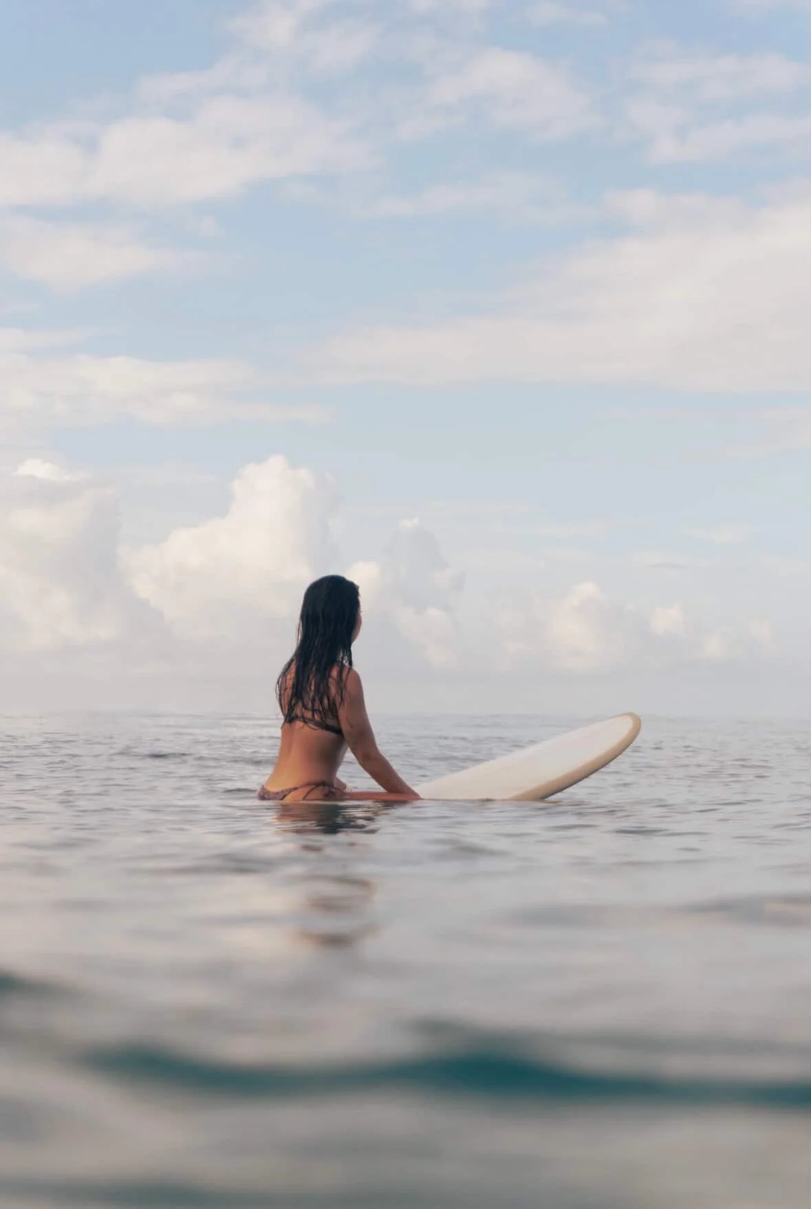 Woman sitting on surfboard in the ocean, looking out at the horizon with a cloudy sky.