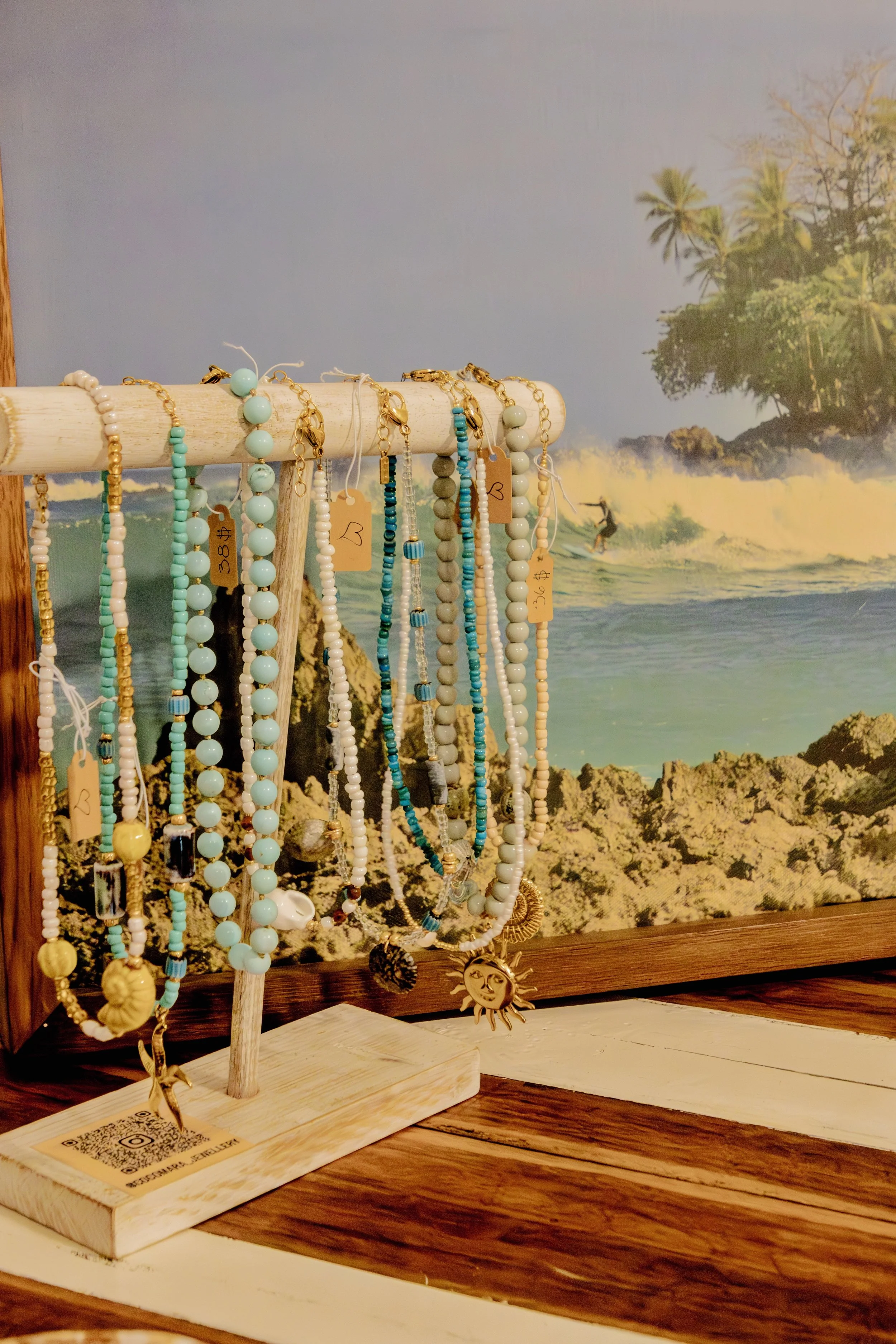 A display of various beaded necklaces on a wooden rack with a beach and ocean scene in the background.